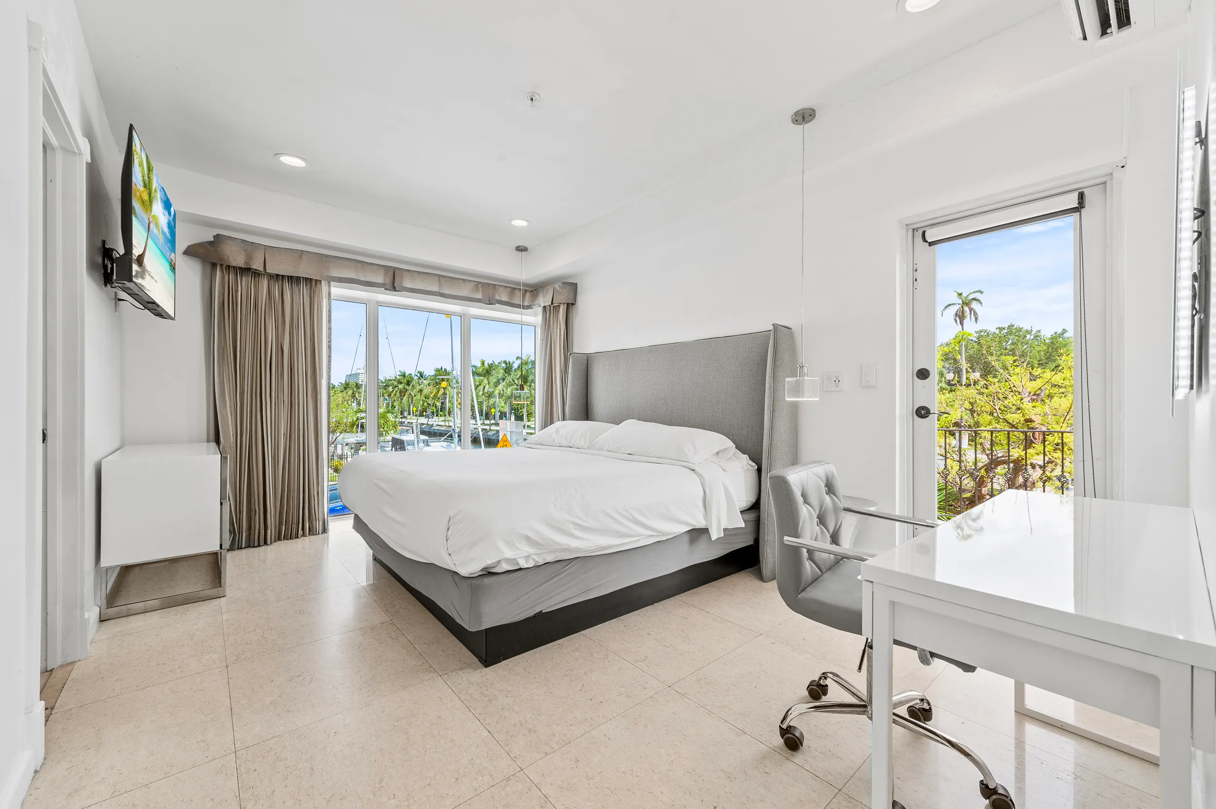 Bright bedroom with a gray upholstered bed, white desk with gray chair, TV on wall, and glass doors showing green trees and boats outside.