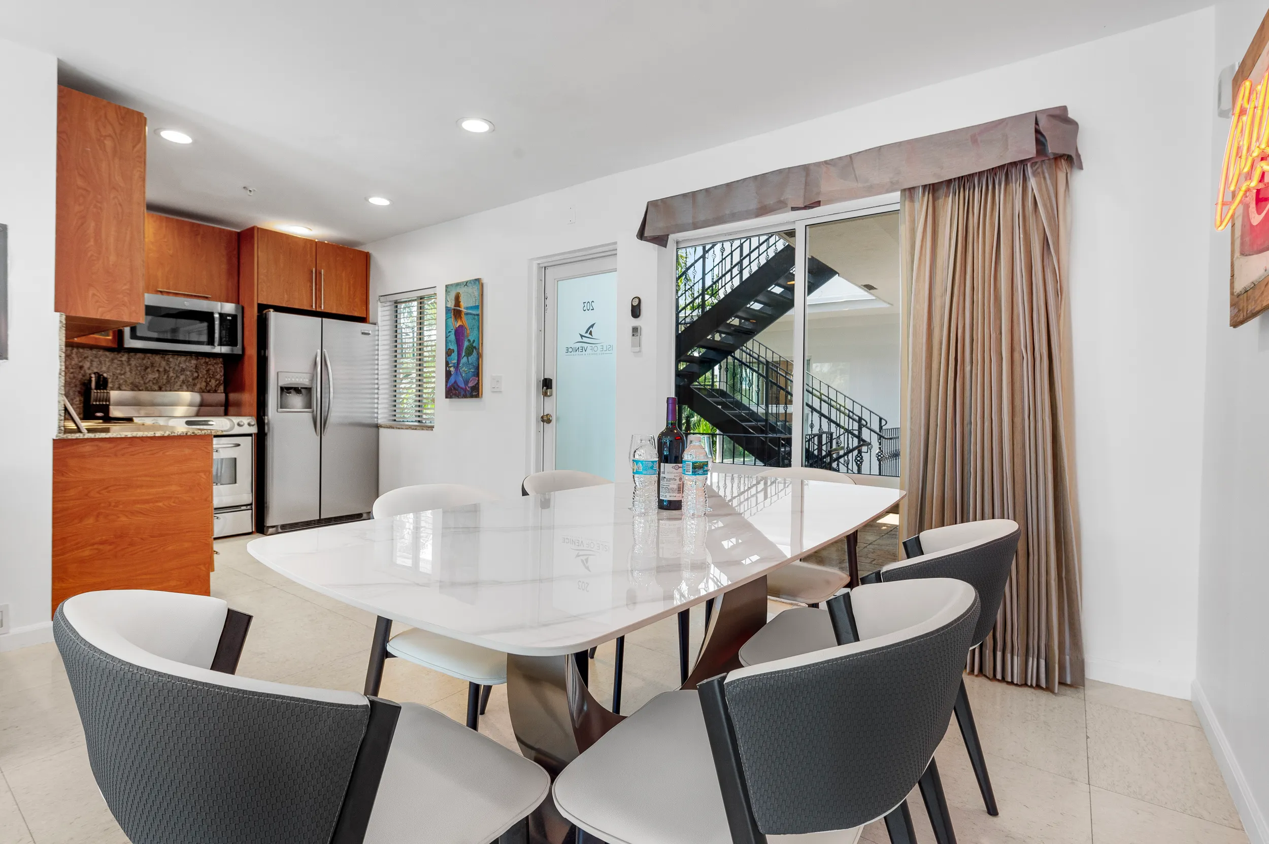 Modern dining area with a marble top table, six gray cushioned chairs, a bottle of wine, two water bottles, and a kitchen with wooden cabinets and stainless steel appliances in the background.