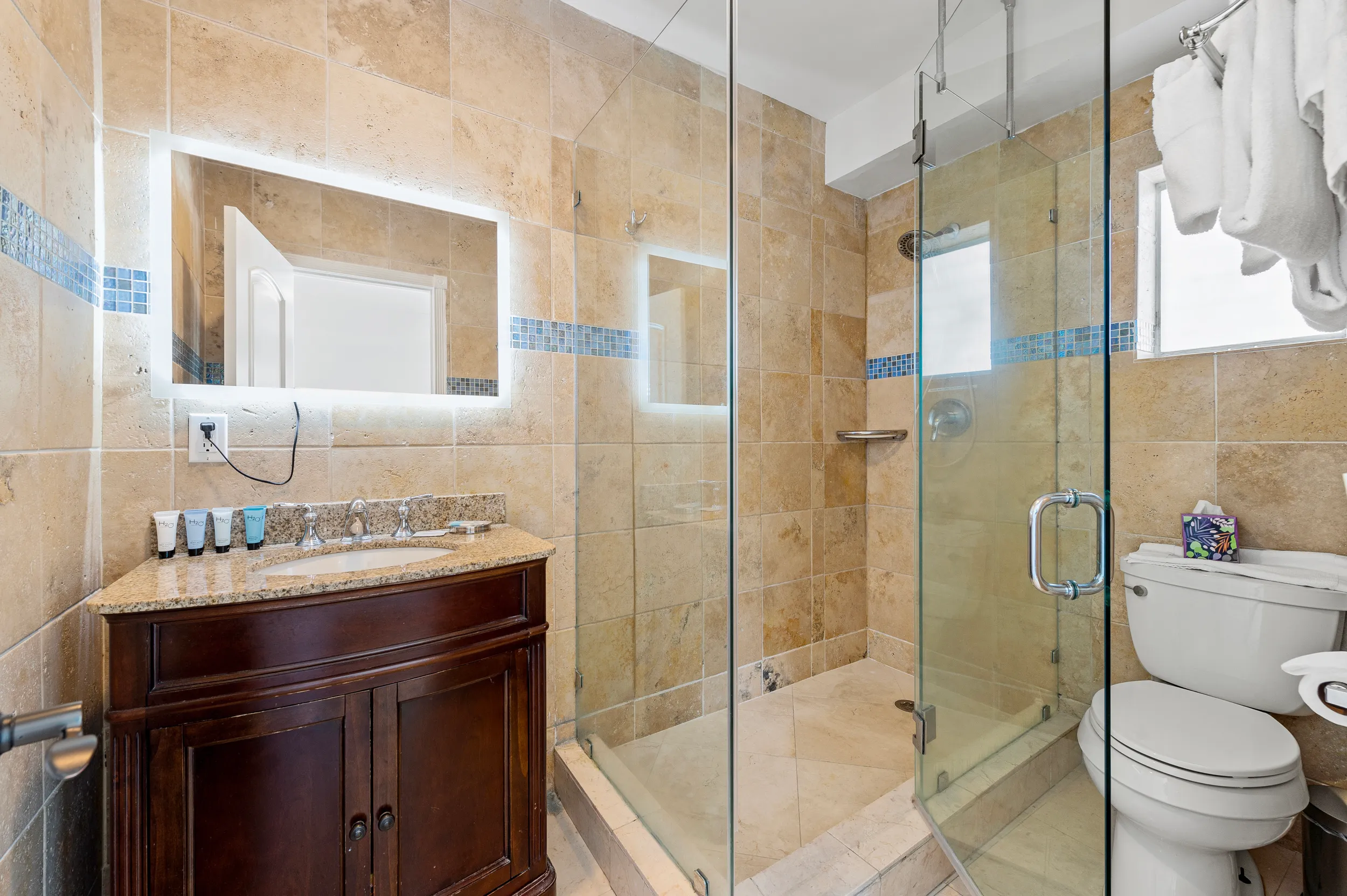 Modern bathroom with beige tiles, glass shower enclosure, dark wood vanity with granite countertop, illuminated mirror, and a white toilet with folded towels.
