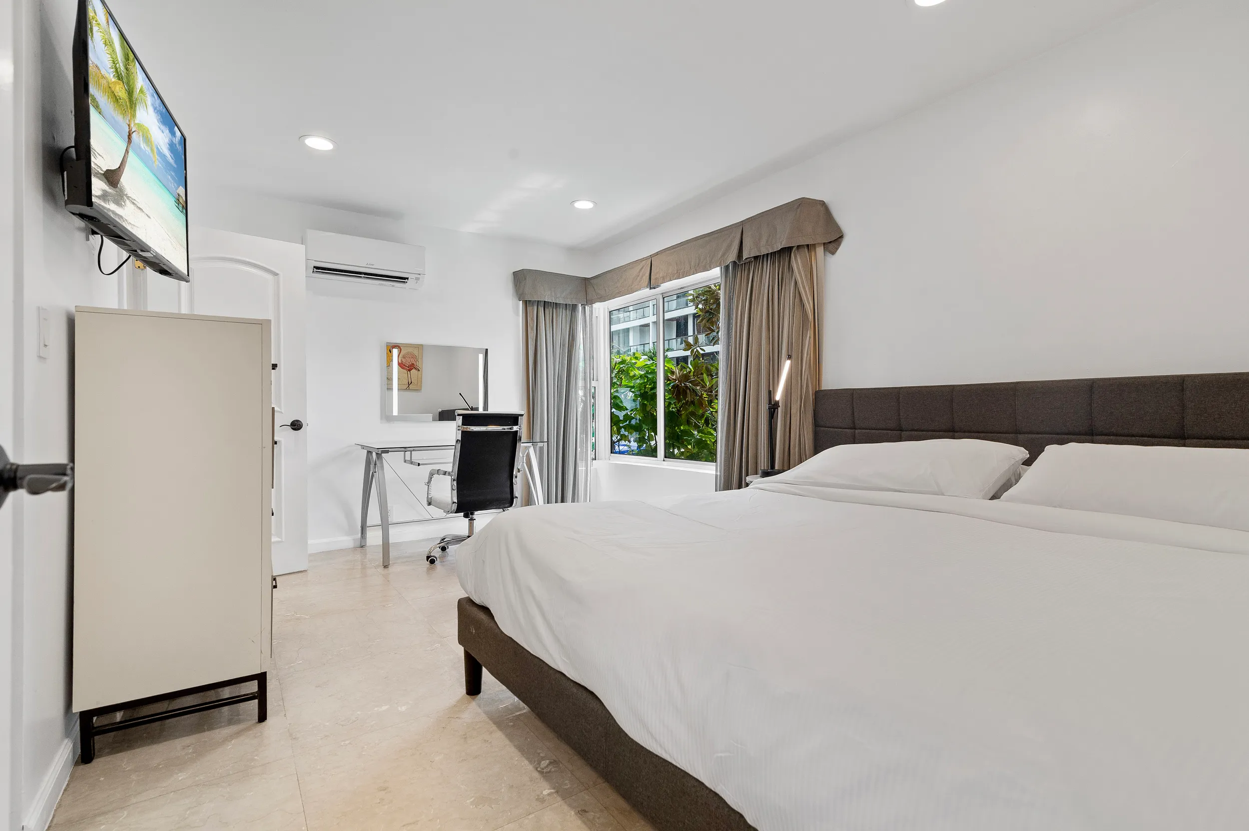 Modern bright bedroom with a king-sized bed, white linens, a work desk with swivel chair, wall-mounted TV, and a large window with beige curtains overlooking greenery.