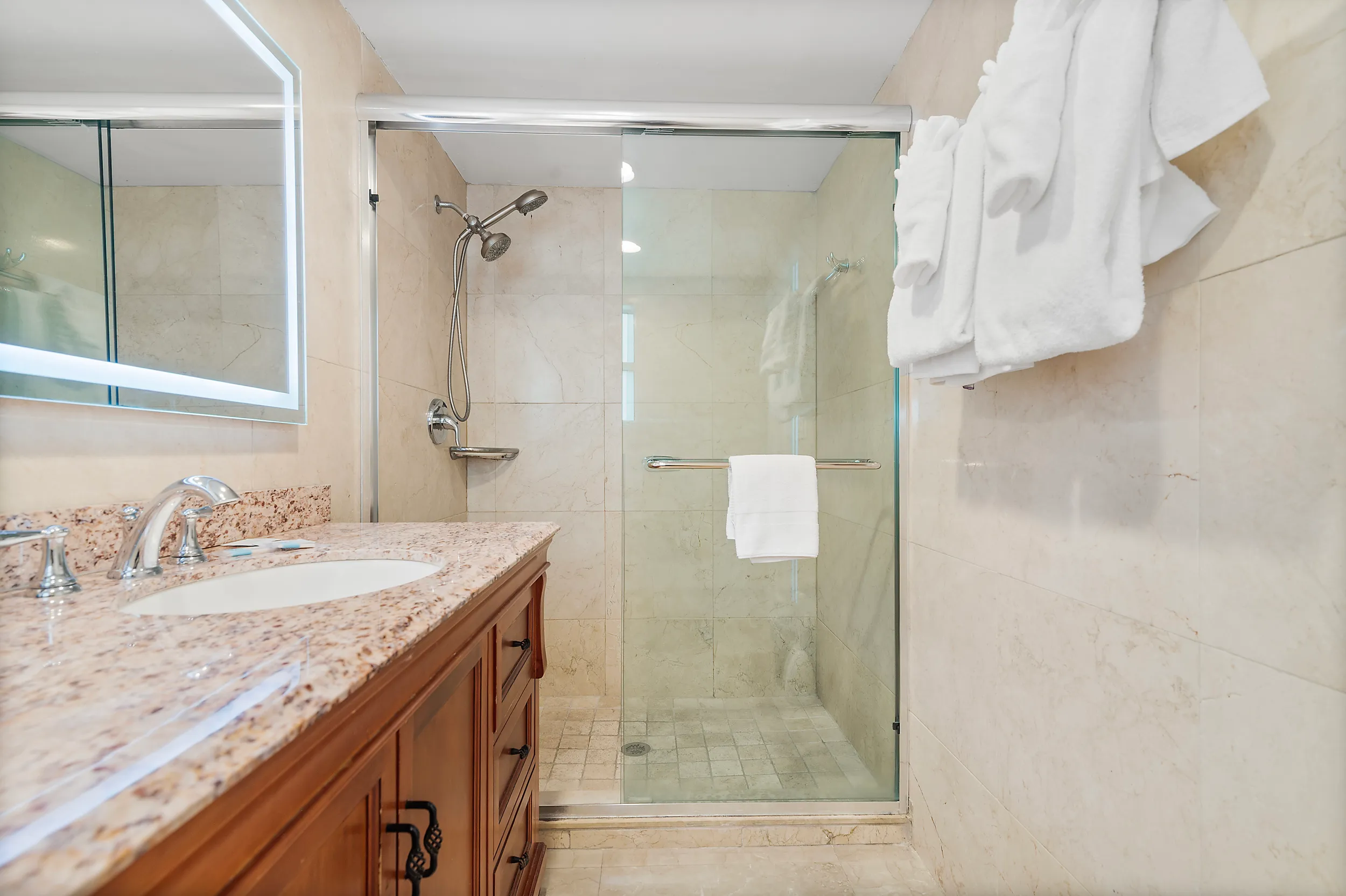 Bathroom with a glass-enclosed walk-in shower, beige tiled walls, wooden vanity with granite countertop, and white towels hanging.