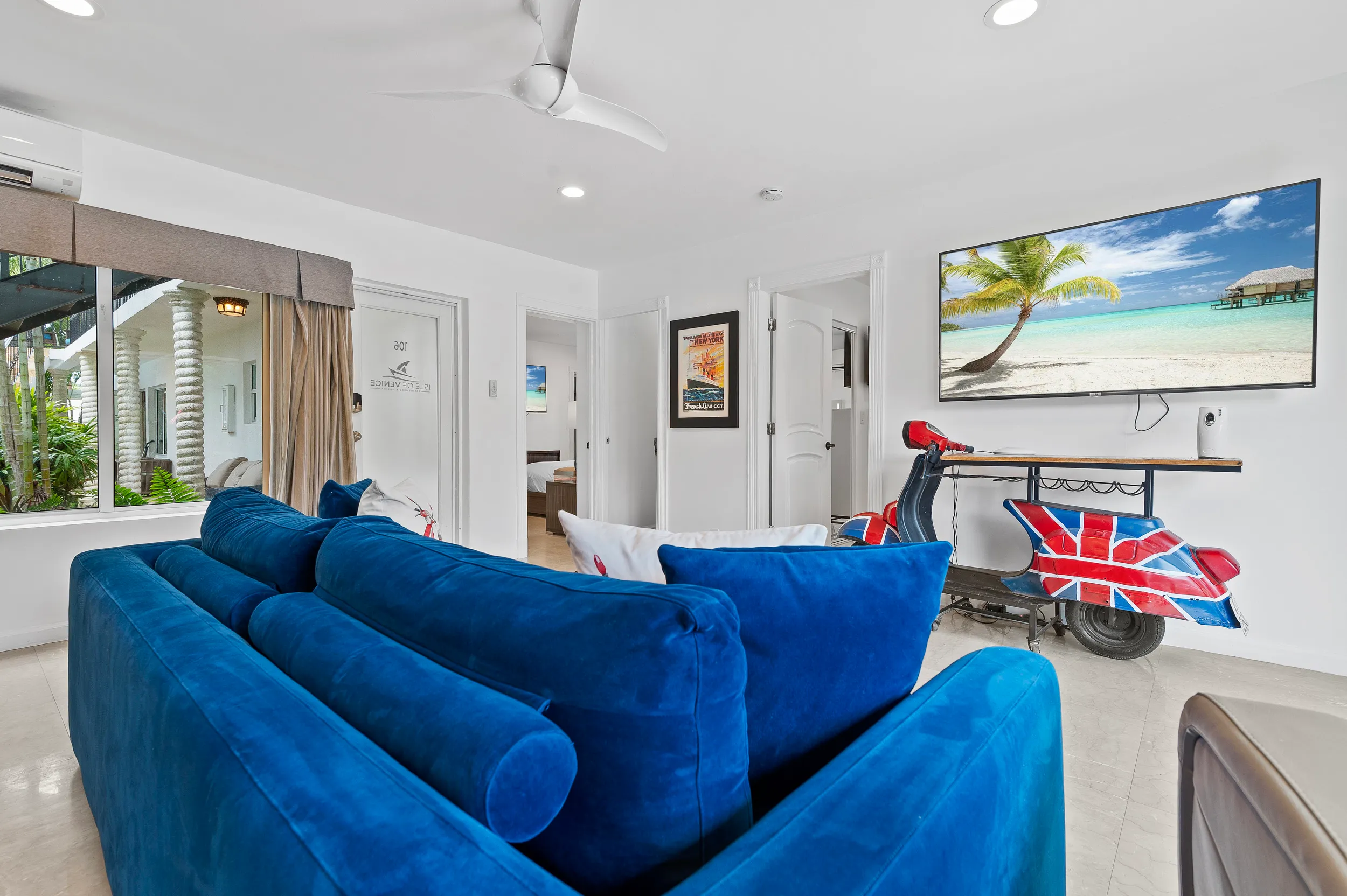 Modern living room with a vibrant blue sofa, large window, ceiling fan, a wall-mounted TV displaying a beach scene, and a scooter-shaped table with a Union Jack design.