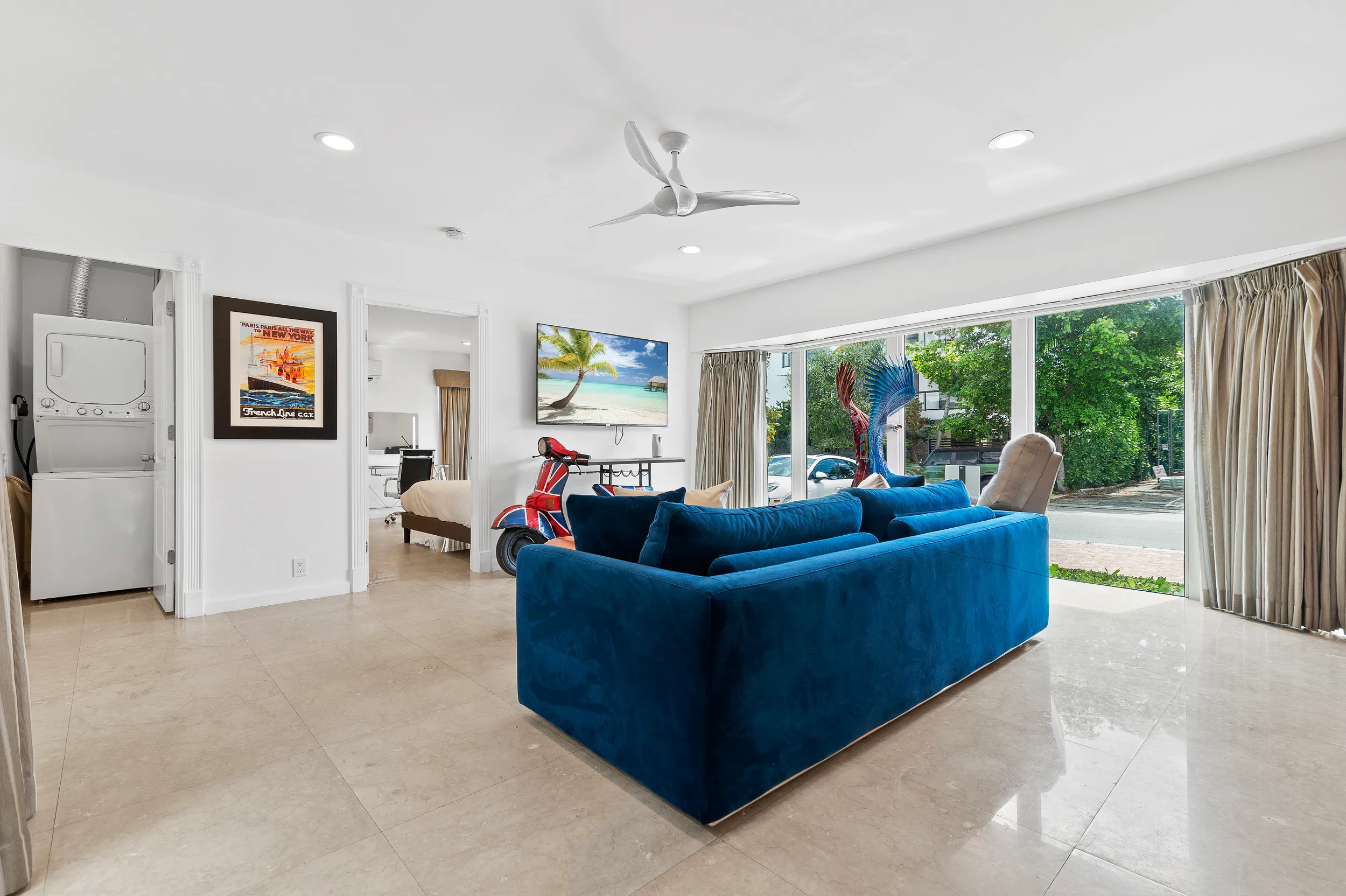 Modern living room with a blue velvet sofa, large windows with beige curtains, a wall-mounted TV displaying a beach scene, a red scooter, and a stacked washer and dryer.