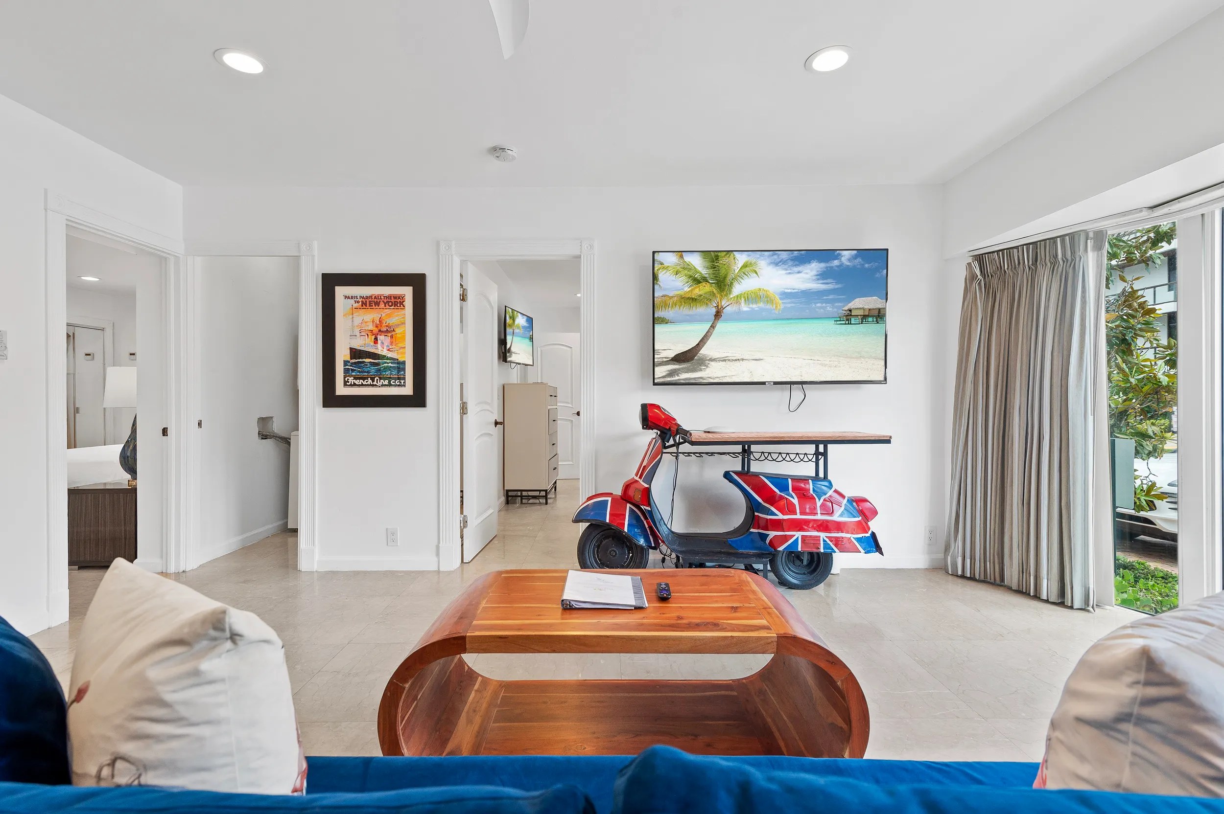Living room with white walls, a wooden coffee table, blue couch cushions, and a unique scooter-shaped bar table with a Union Jack design under a wall-mounted TV showing a beach scene.