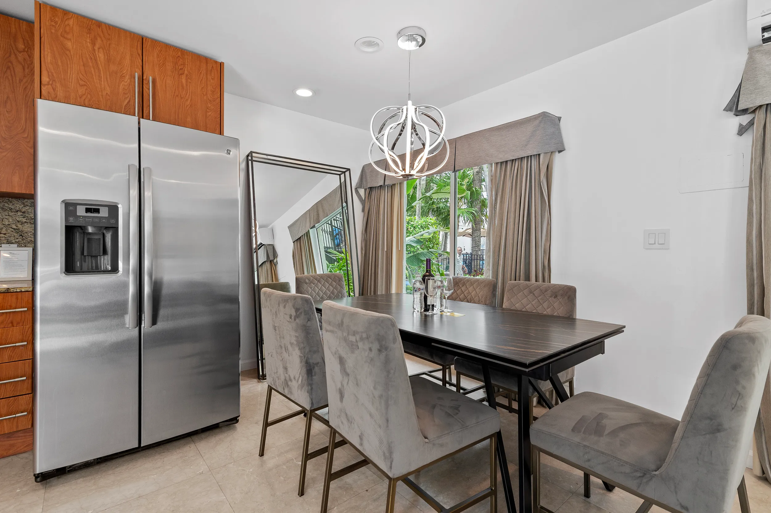 Modern dining area with a dark wood table, six gray upholstered chairs, a stainless steel refrigerator, large standing mirror, and a window with beige curtains showing green foliage outside.