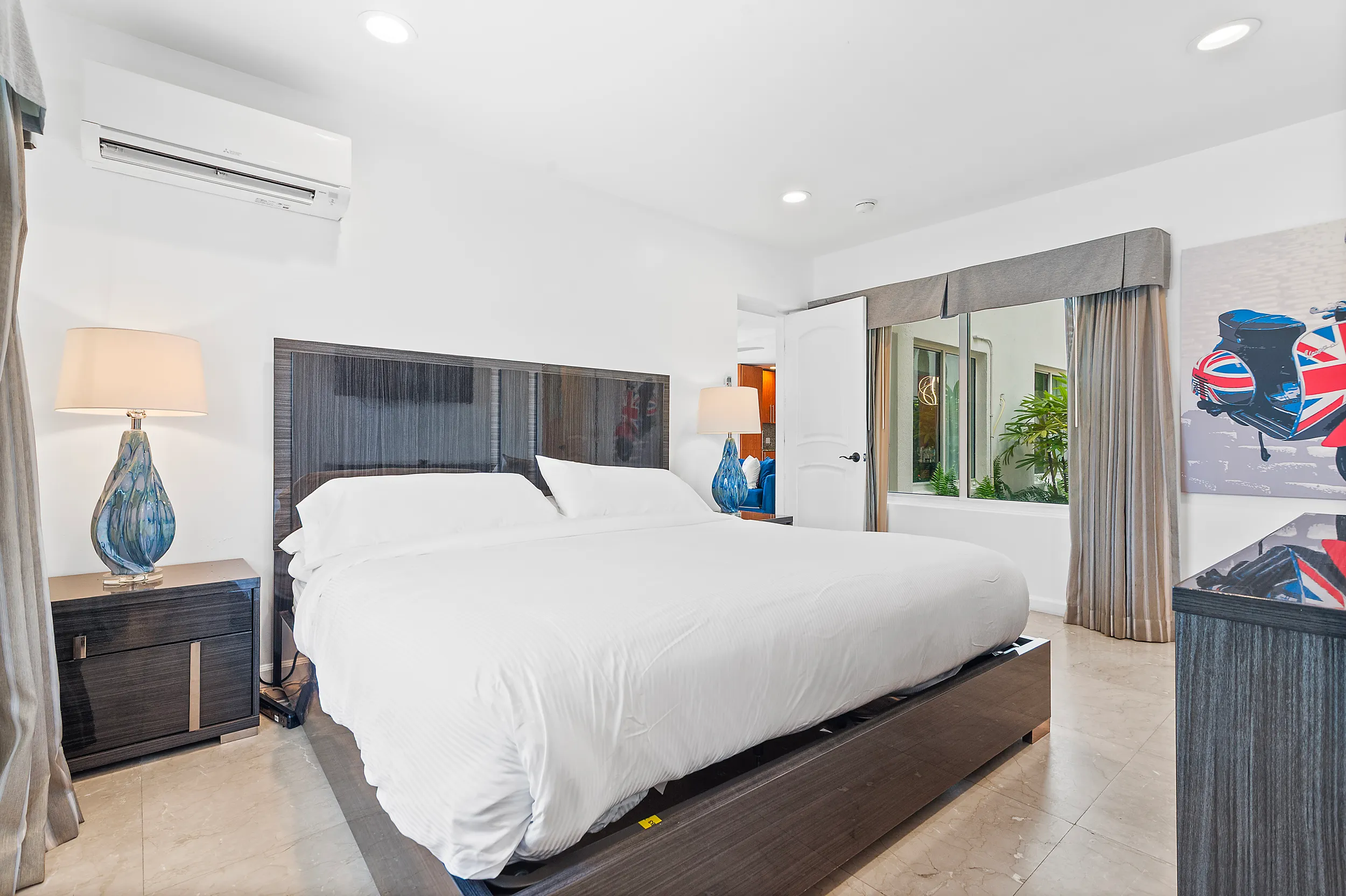 Modern bedroom with a king-sized bed, white bedding, two blue glass lamps on dark wood nightstands, and a window with beige curtains.