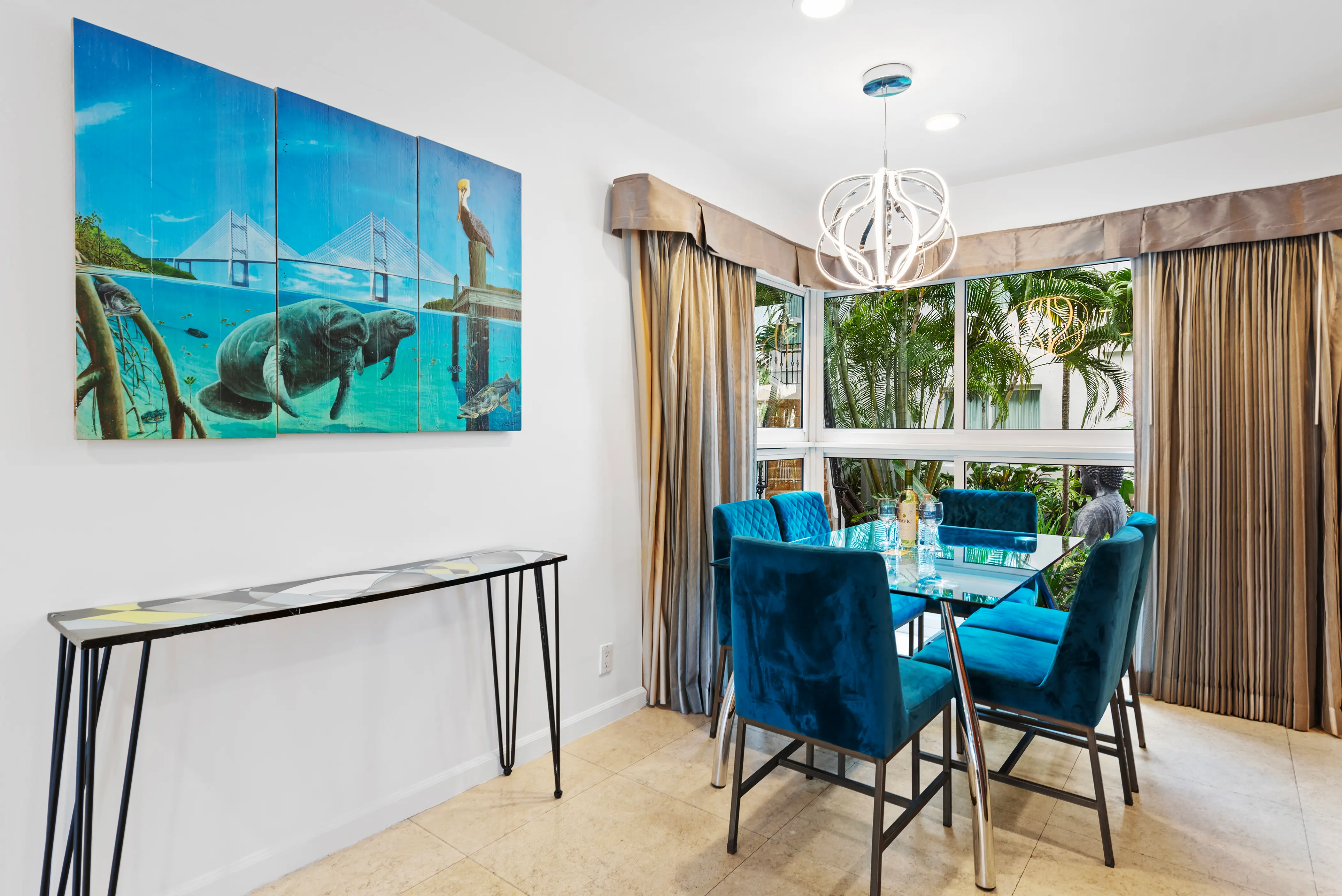 Modern dining area with a glass table surrounded by six blue velvet chairs, large windows with beige curtains, and a wall painting of manatees and a bridge.