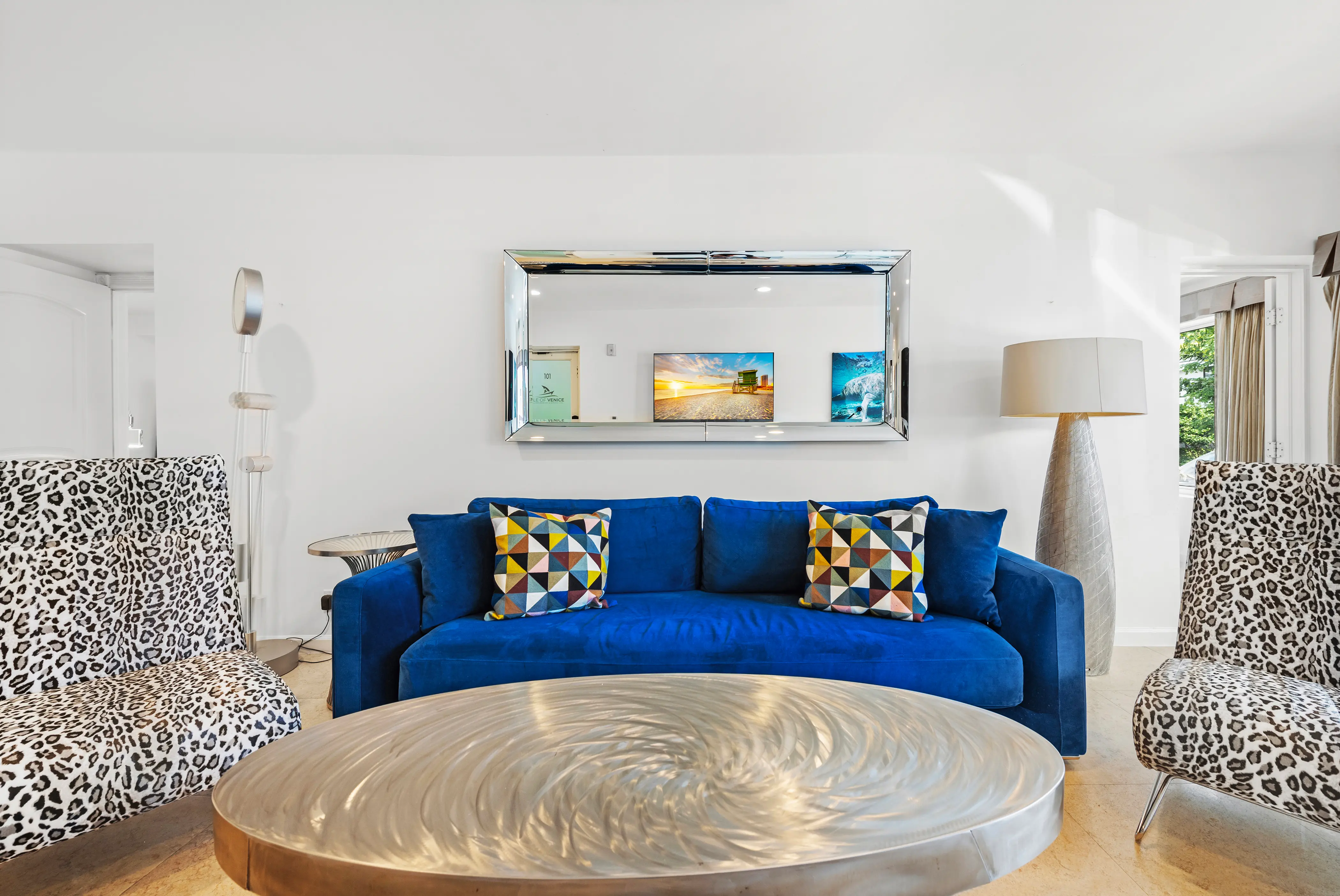 Living room with a blue sofa, two leopard-print chairs, a round metallic coffee table, and a large rectangular mirror on a white wall.