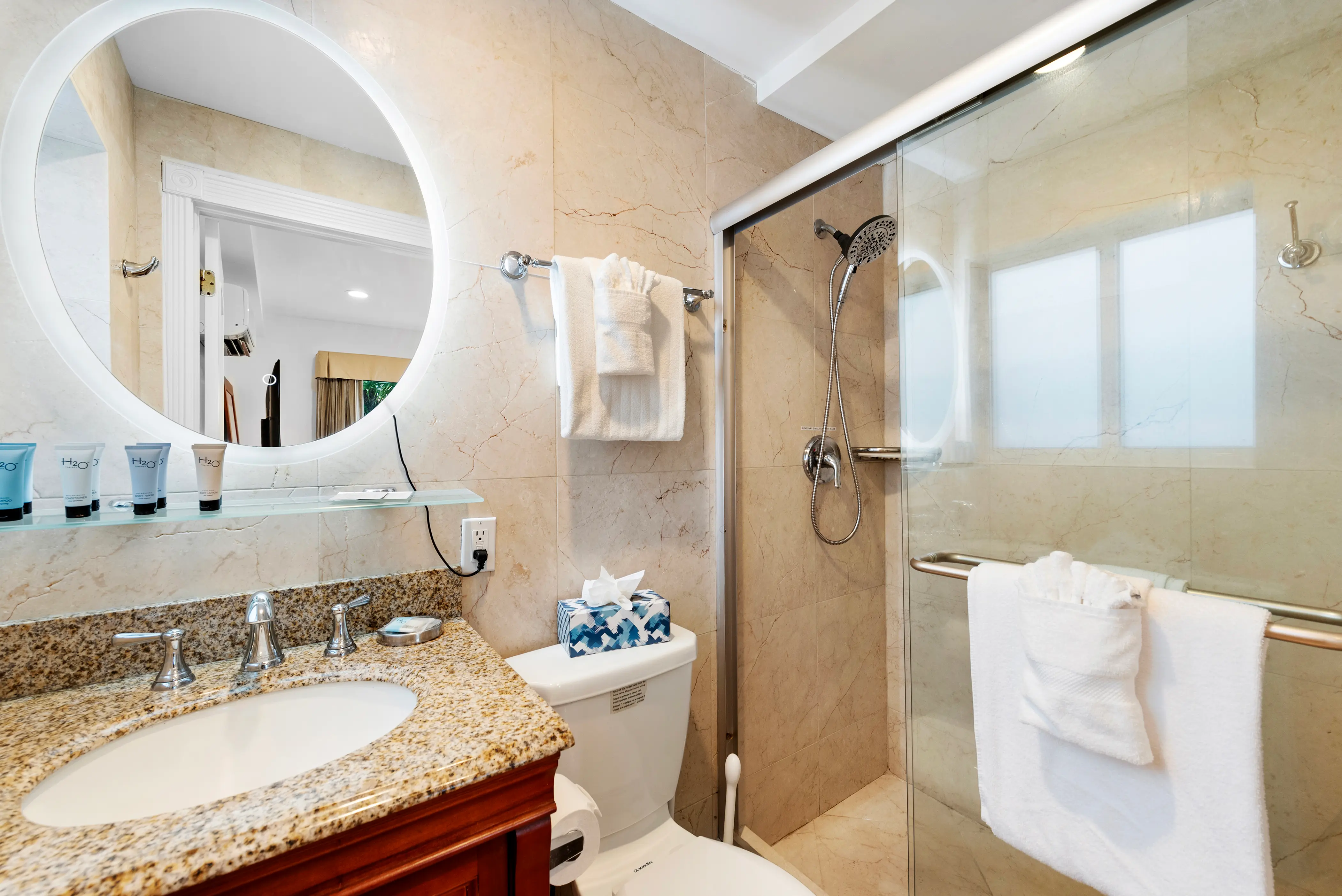 Modern bathroom with granite countertop, round illuminated mirror, toiletries, toilet with tissue box, and glass-enclosed shower with towels.