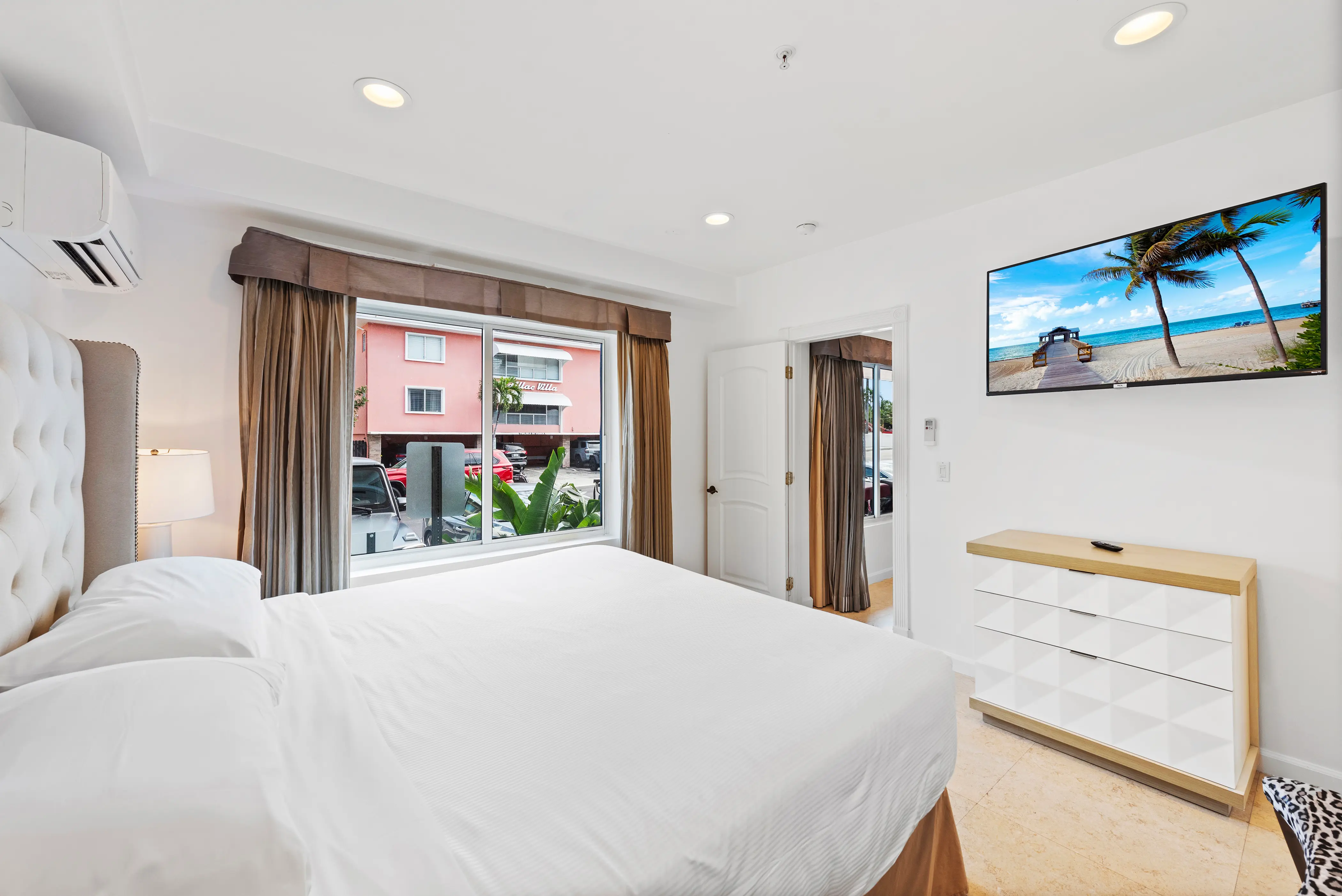 Bright hotel room with white bed, large window with beige curtains, wall-mounted TV showing beach scene, and modern dresser.