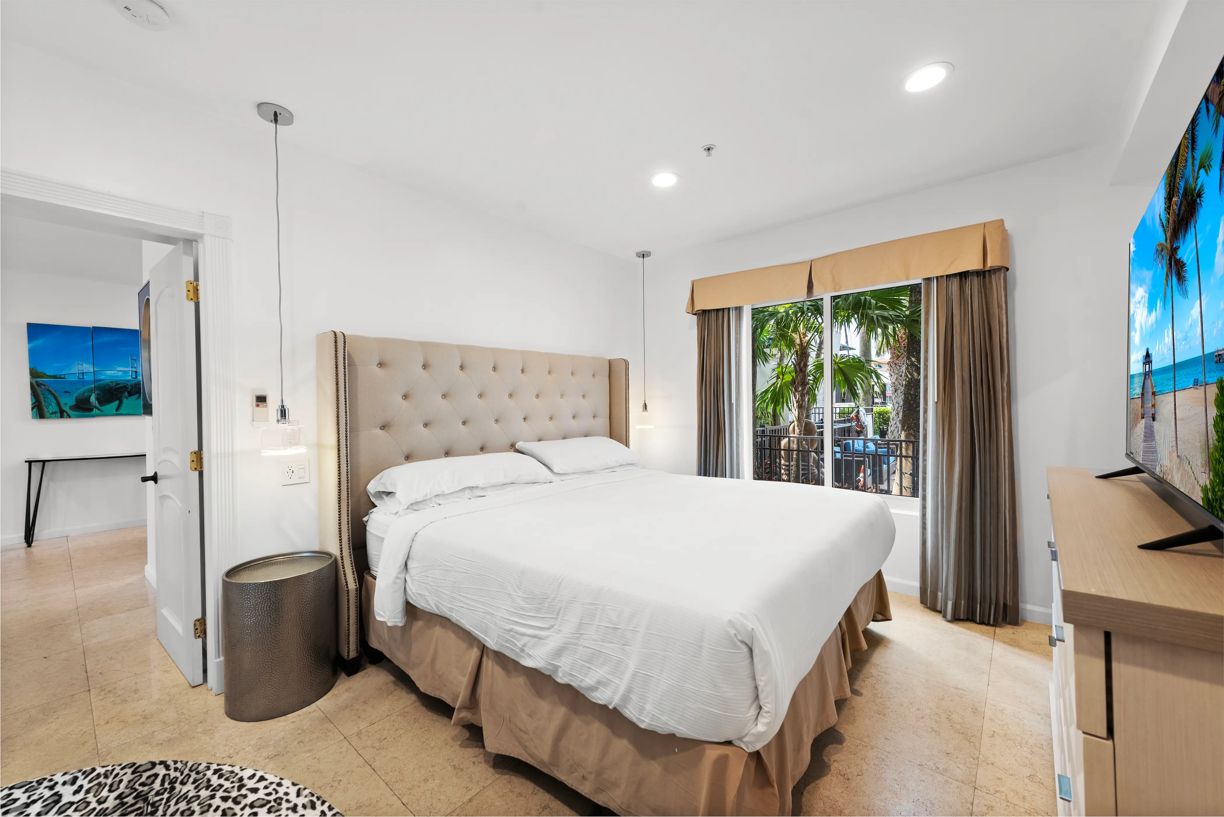 Bright hotel bedroom with a large beige tufted headboard bed, white linens, window with palm trees outside, and a TV showing a beach scene.