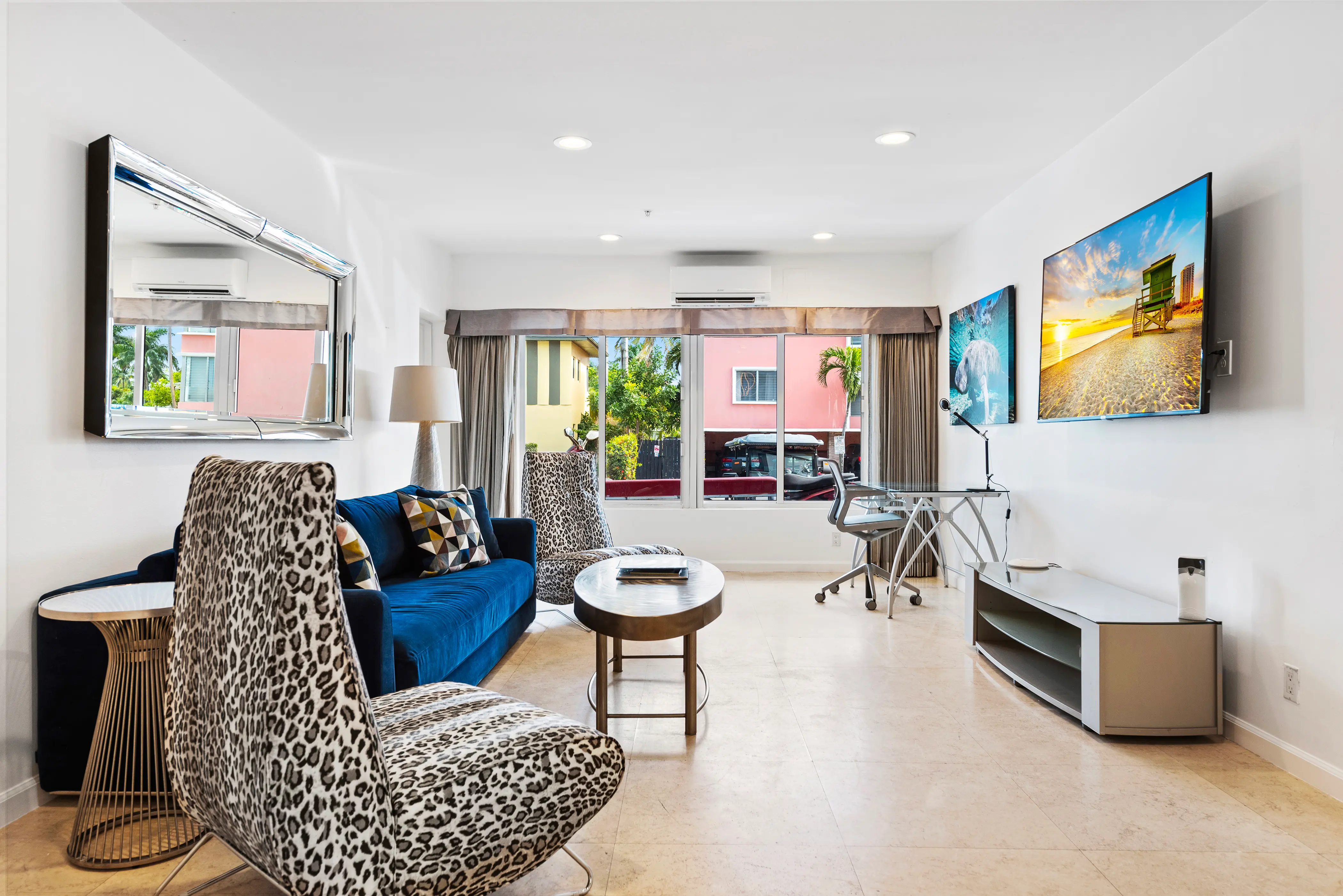 Modern living room with leopard print chairs, a blue sofa, glass desk with rolling chair, and two wall-mounted TVs.