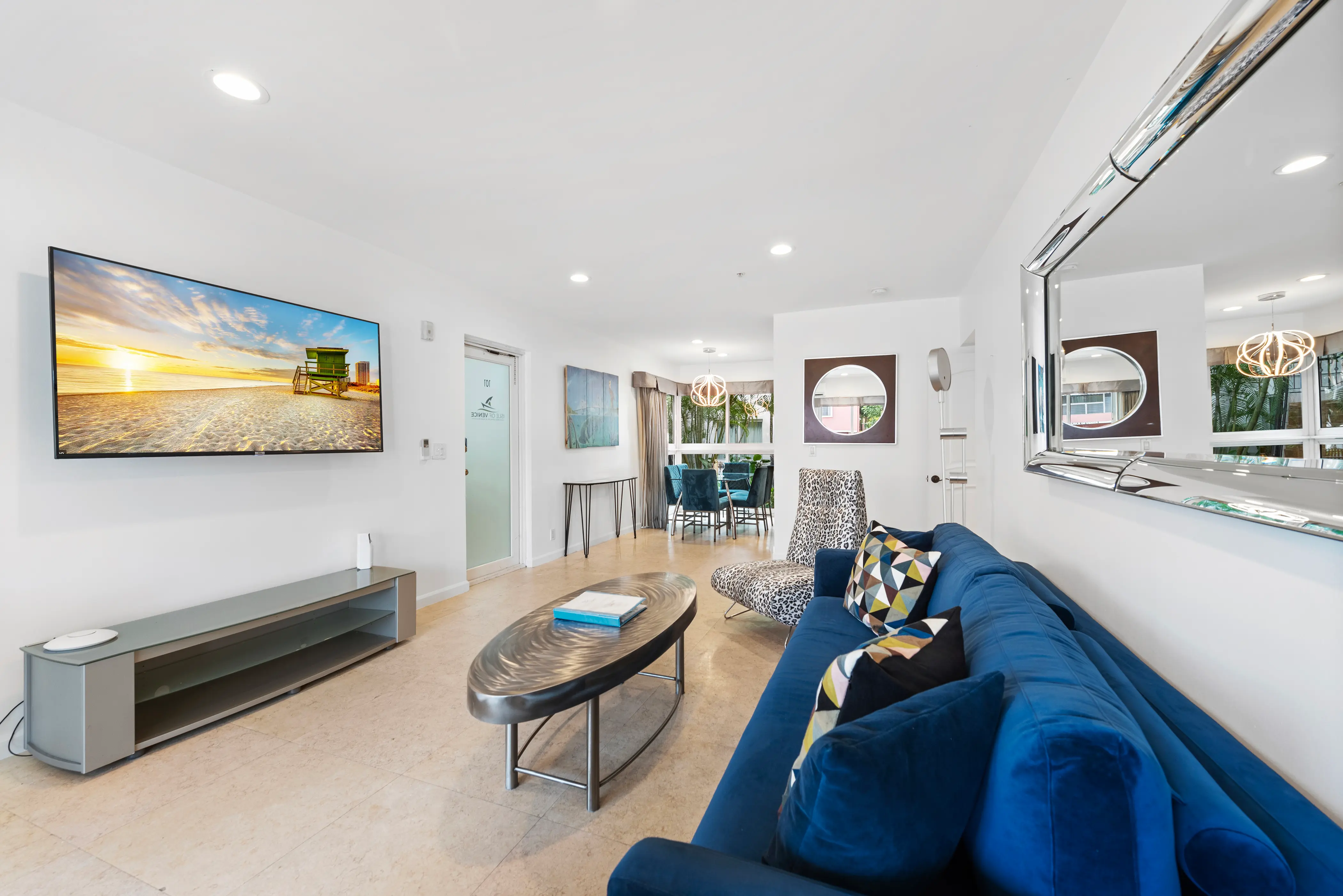 Modern living room with blue velvet sofa, leopard print chair, oval metallic coffee table, wall-mounted TV showing a beach sunset, and a dining area in the background.