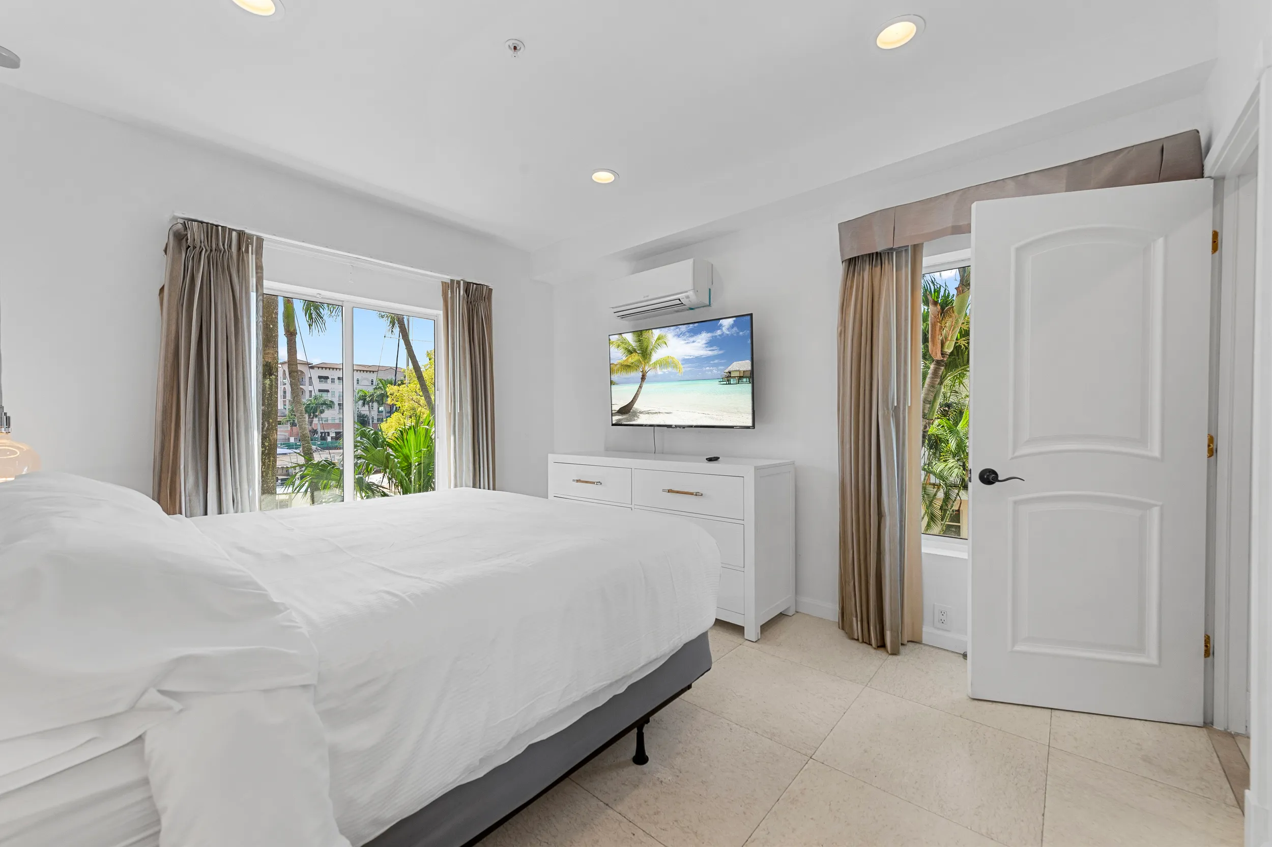 Bright bedroom with white bed linens, large windows with beige curtains, a wall-mounted TV showing a tropical beach scene, and a white dresser.