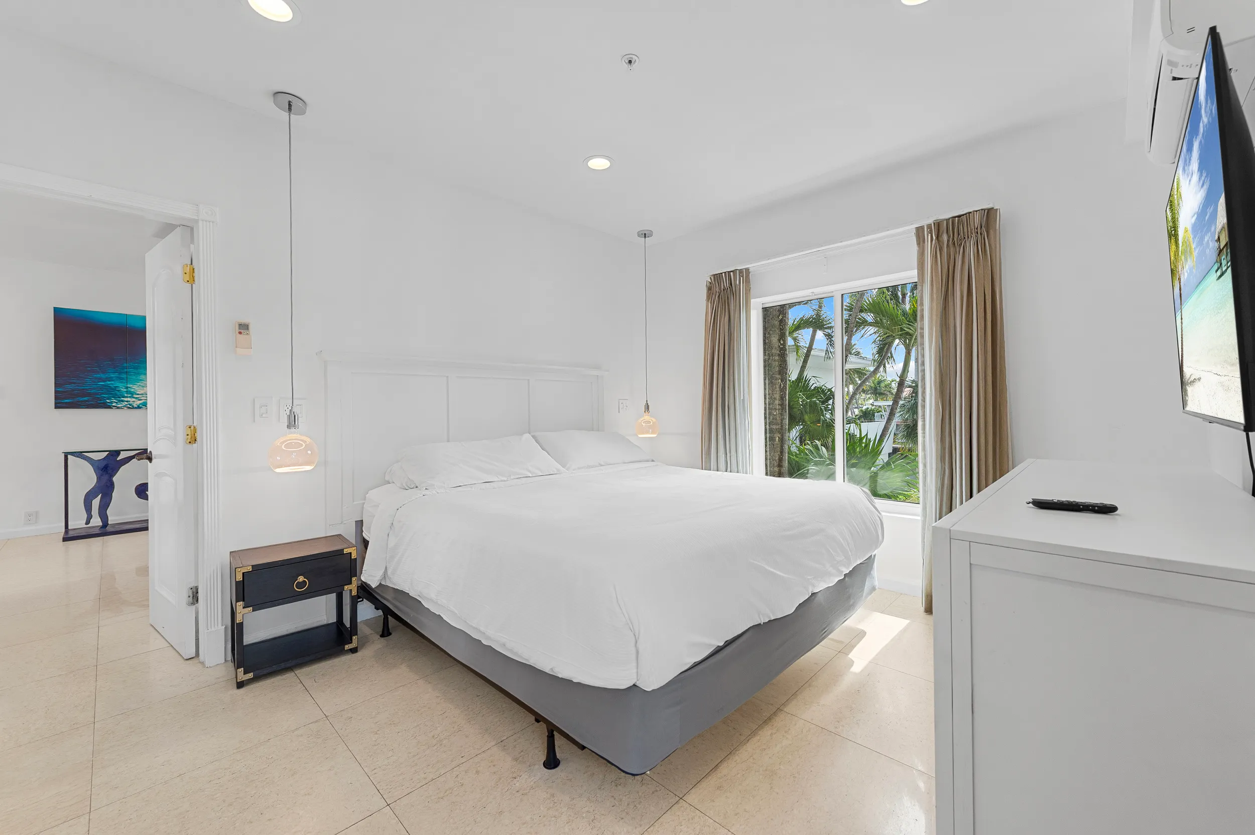 Bright bedroom with a large bed with white bedding, a black bedside table, hanging pendant lights, a window with beige curtains showing palm trees outside, and a TV mounted on the wall.