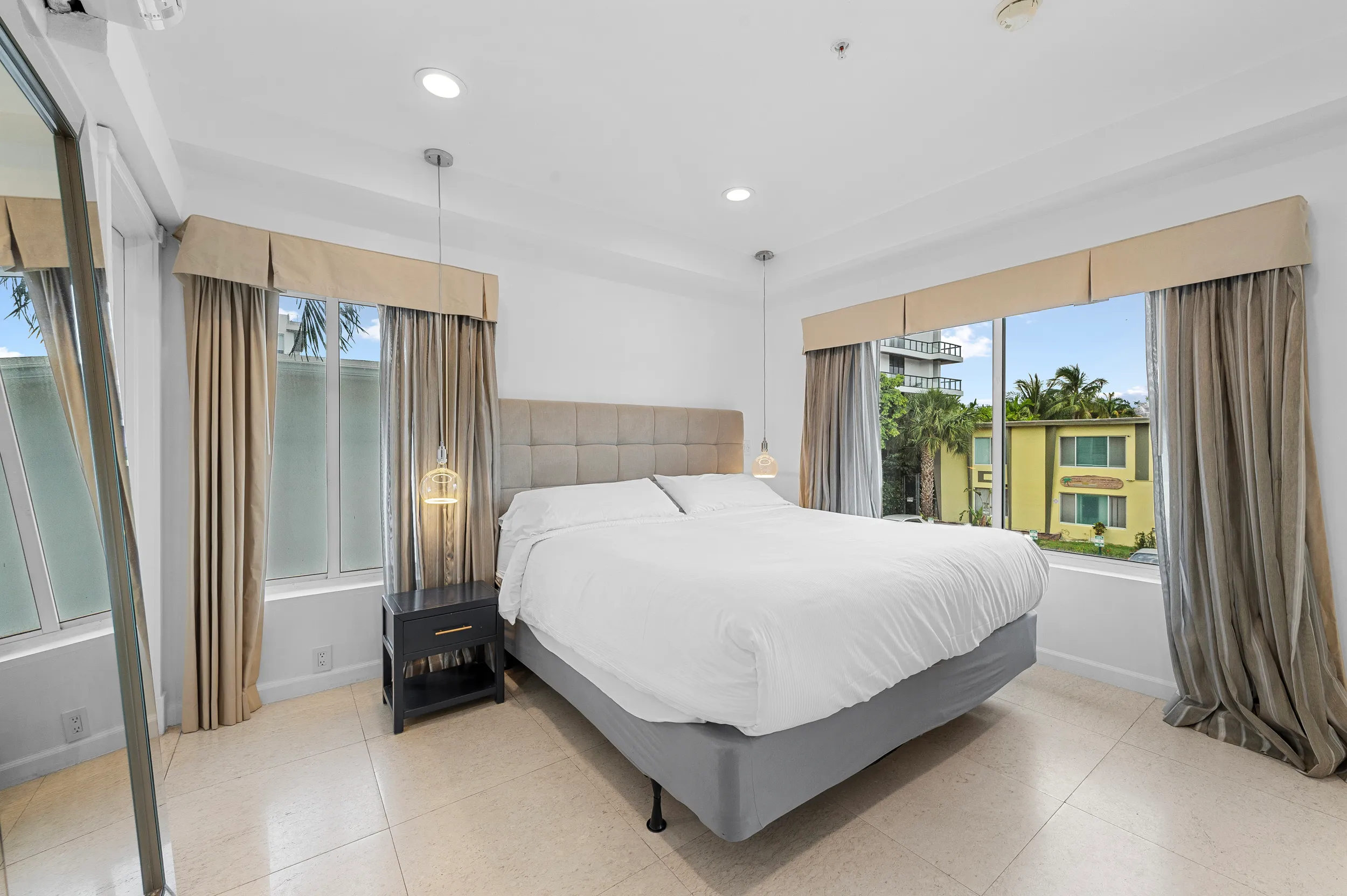 Bright bedroom with a large bed, beige headboard, white bedding, two hanging bedside lamps, and large windows with beige curtains showing a view of buildings and palm trees.
