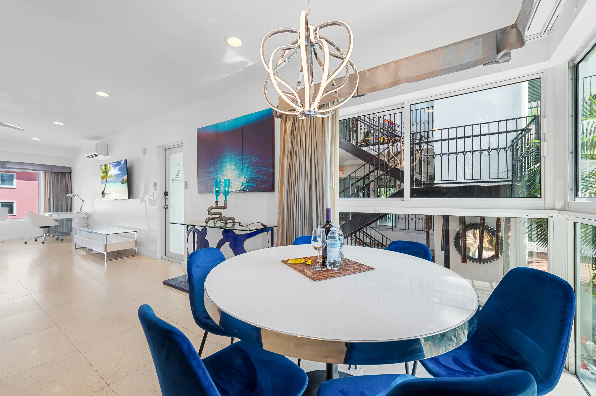 Modern dining area with round white table, blue velvet chairs, a contemporary chandelier, and large windows showing an outdoor staircase.