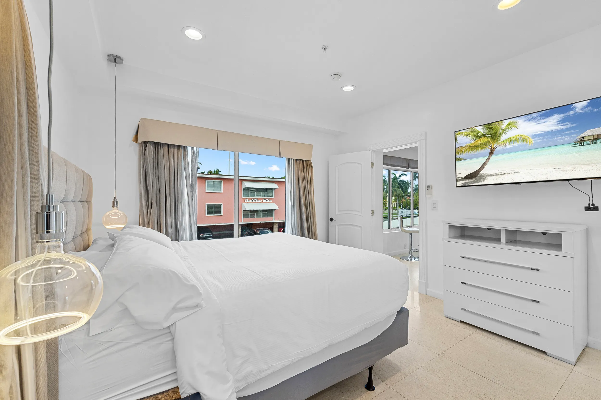 Bright bedroom with a large white bed, pendant lights, a window showing a building outside, and a TV displaying a tropical beach scene.