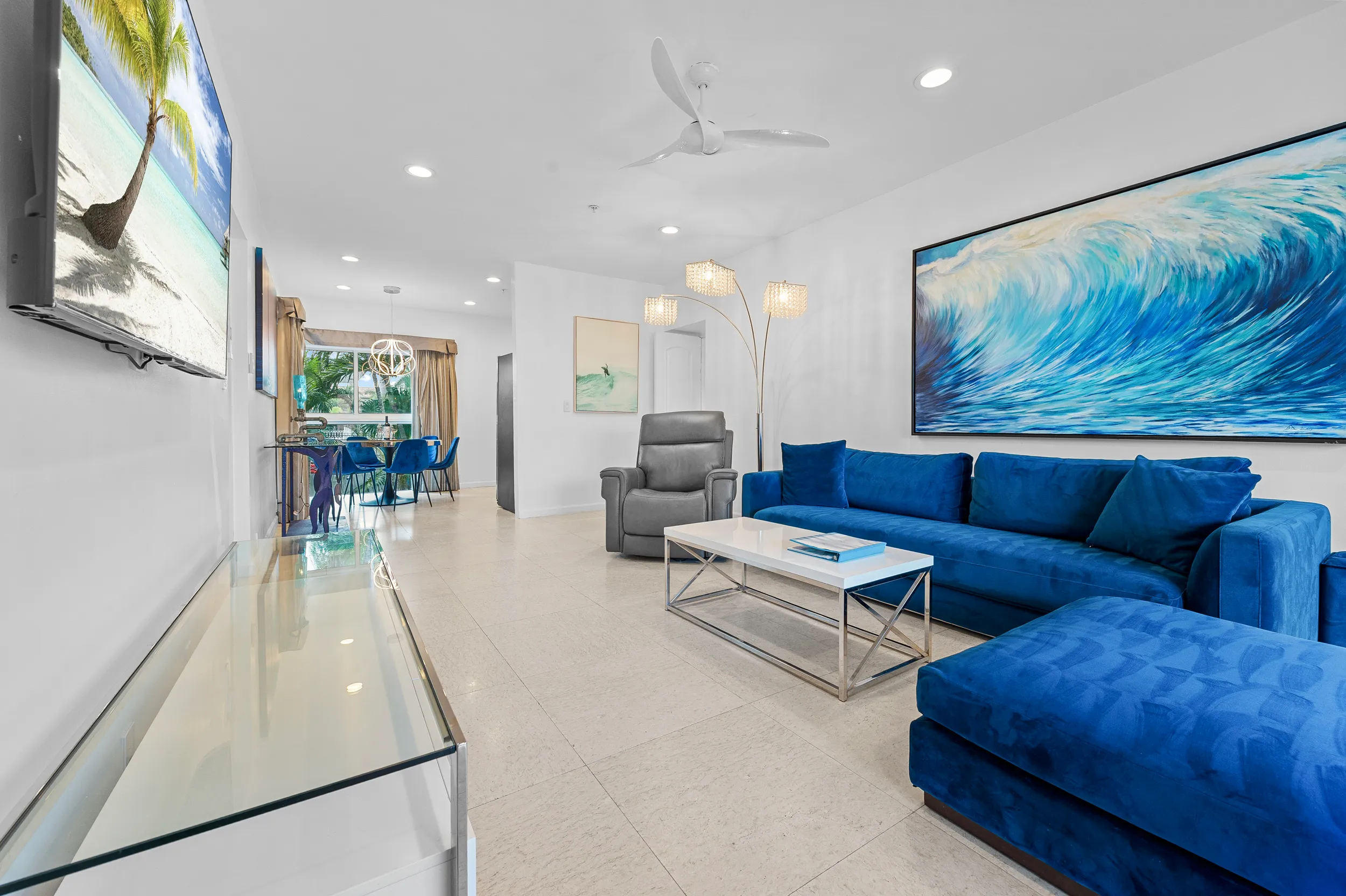 Modern living room with a blue velvet sectional sofa, a gray armchair, a white coffee table, and ocean-themed artwork on the walls.