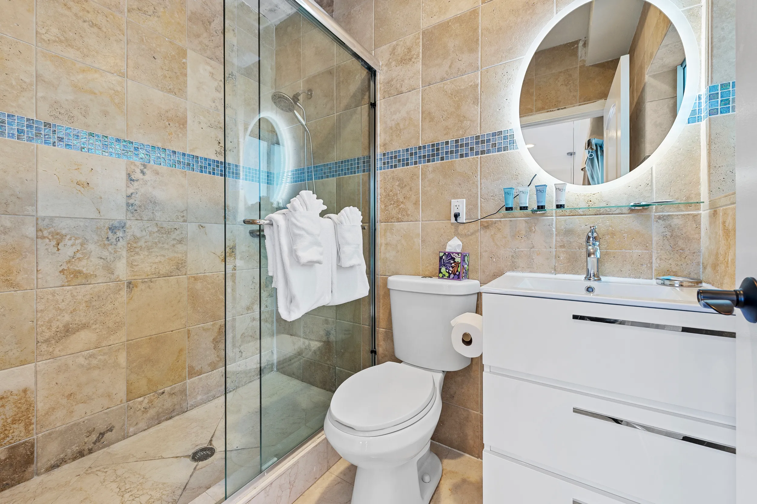 Modern bathroom with beige tiled walls, glass shower enclosure, white toilet, illuminated round mirror, and white vanity cabinet.