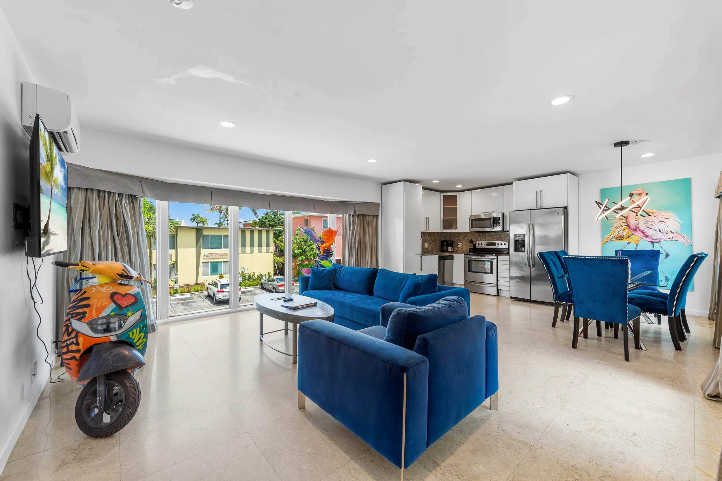 Modern open living space with blue velvet sofa set, dining area with six blue chairs, stainless steel kitchen appliances, and graffiti-decorated orange scooter.