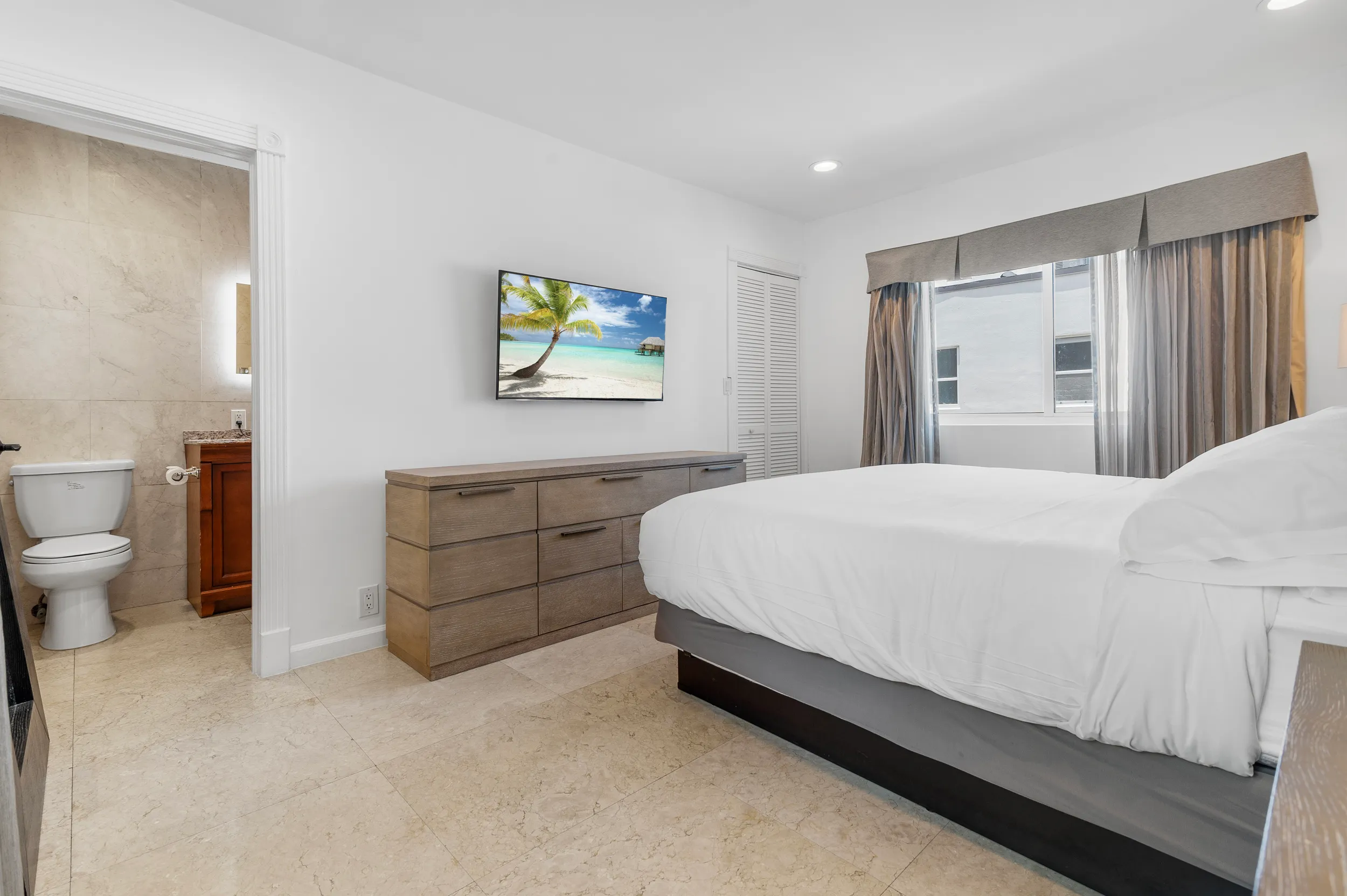 Modern bedroom with white bedding, wooden dresser, wall-mounted TV showing beach scene, and a bathroom partially visible.
