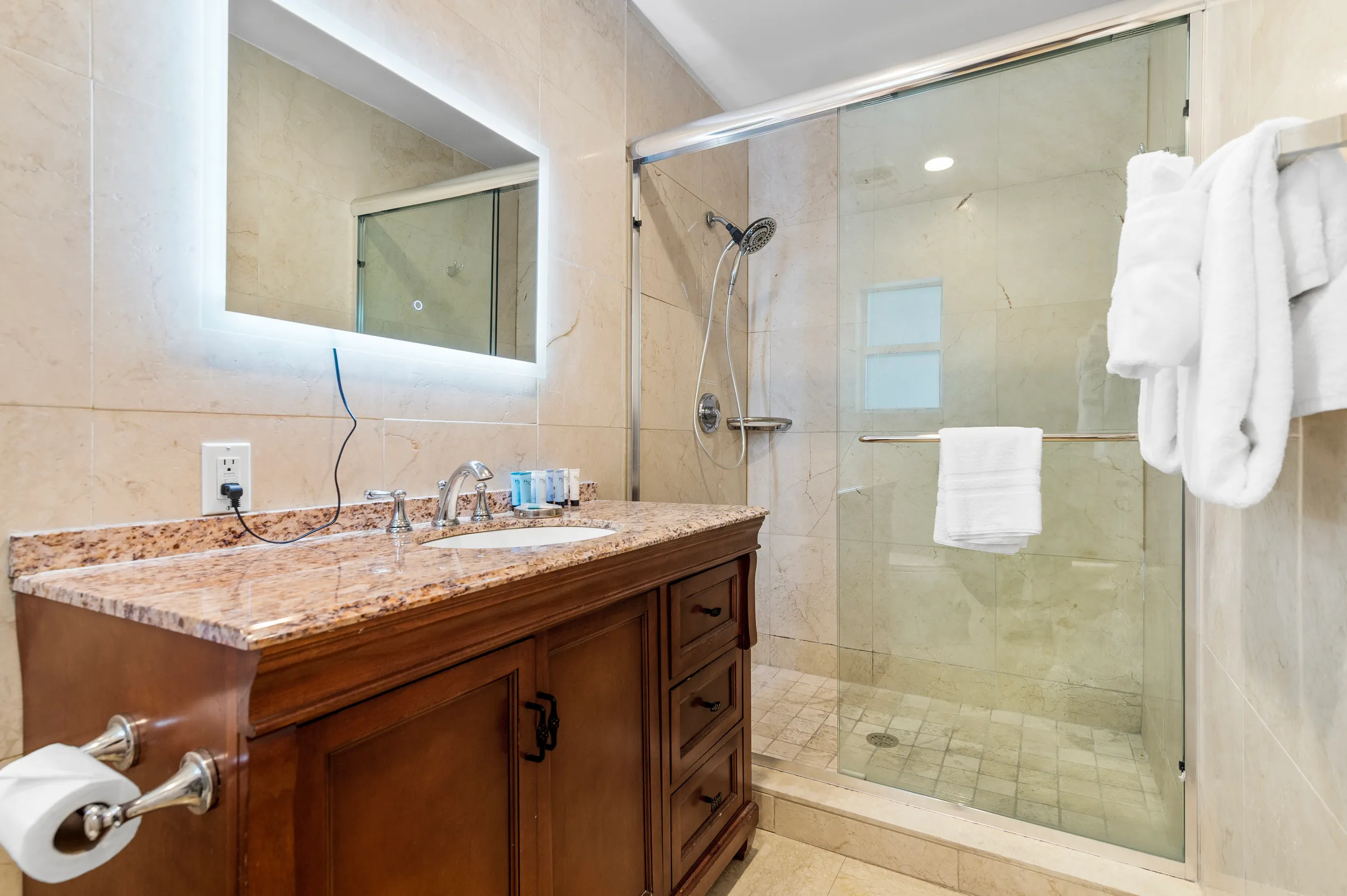 Modern bathroom with a glass-enclosed shower, granite countertop vanity with sink, illuminated mirror, and white towels.