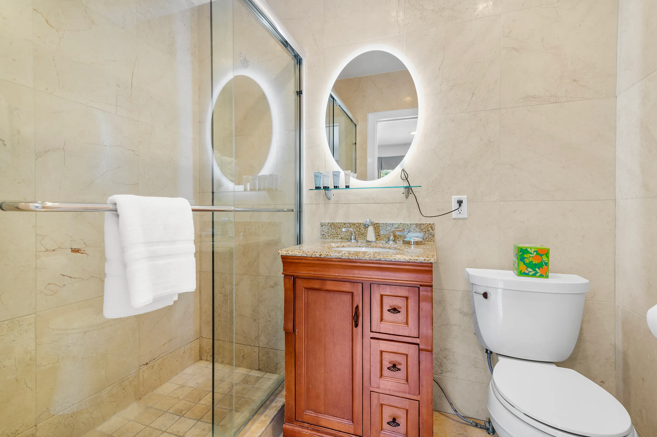 Bathroom with a wooden vanity topped with granite, an illuminated oval mirror, glass shower enclosure with a towel bar holding white towels, and a white toilet with a colorful tissue box on the tank.