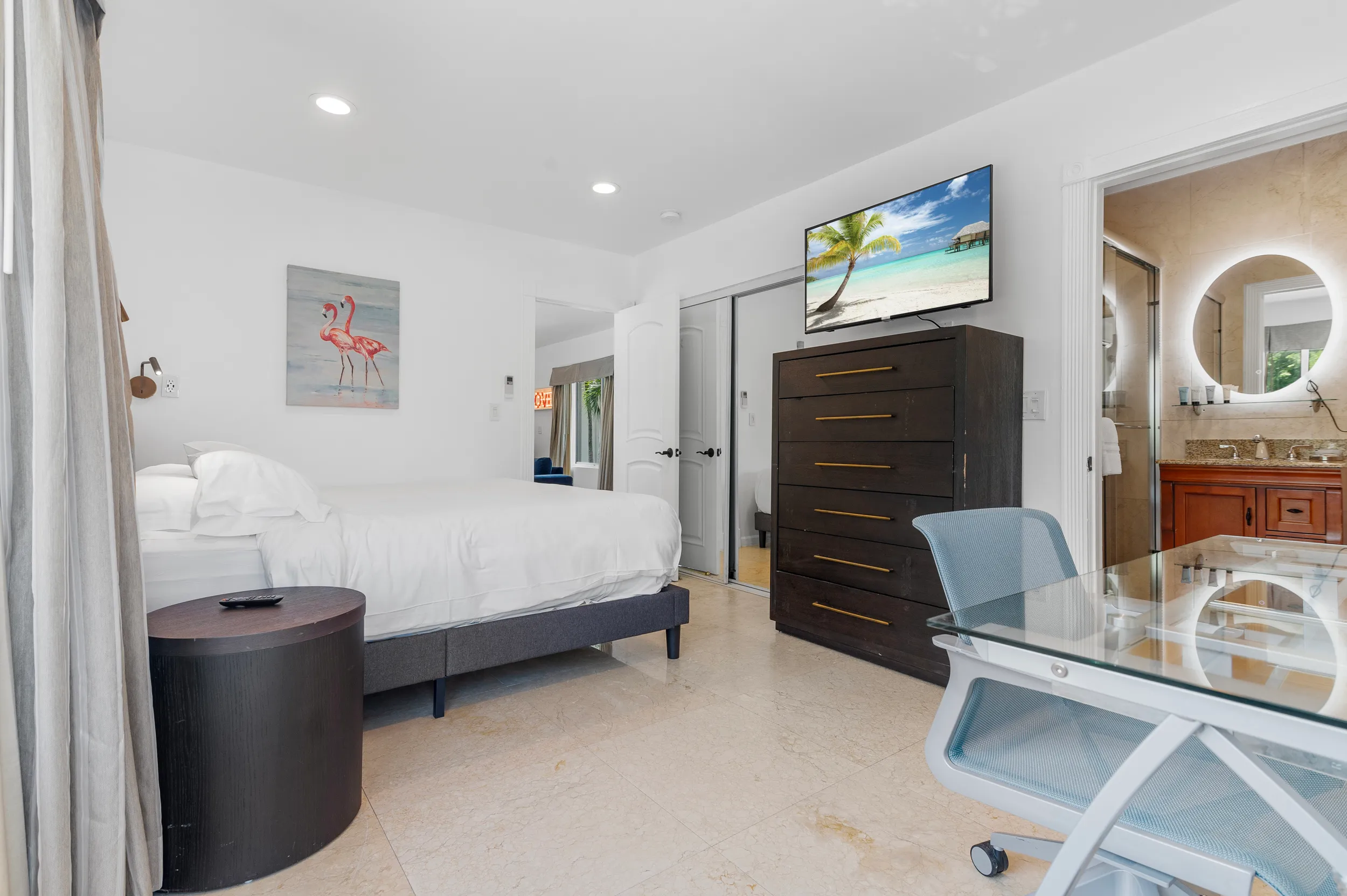 Bright bedroom with a white bed, flamingo wall art, dark wooden dresser with a beach scene on TV, glass desk with blue chair, and an open door to a bathroom with a round illuminated mirror.