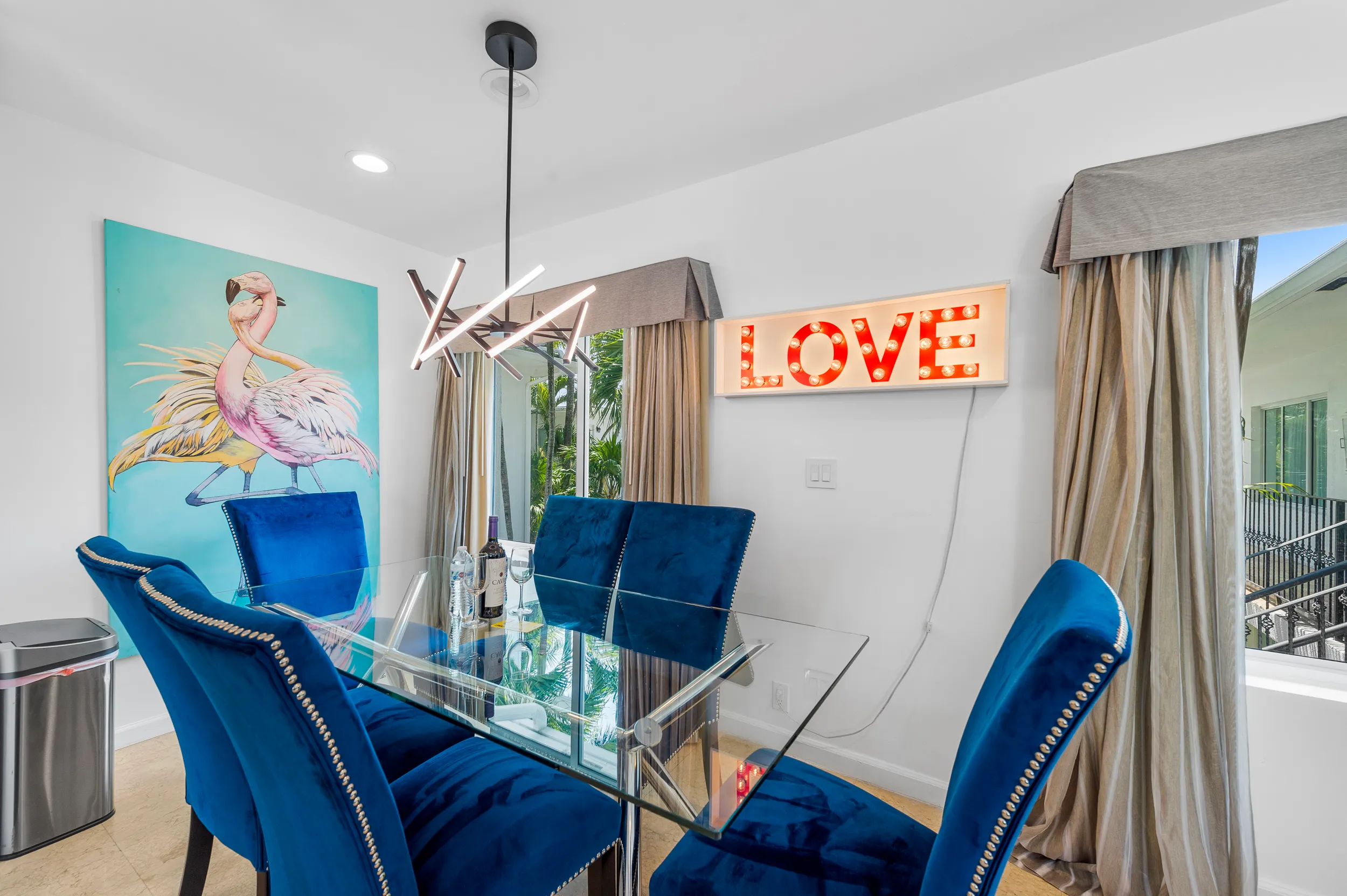 Modern dining room with a glass table, six blue velvet chairs, flamingo artwork, and illuminated LOVE sign on the wall.