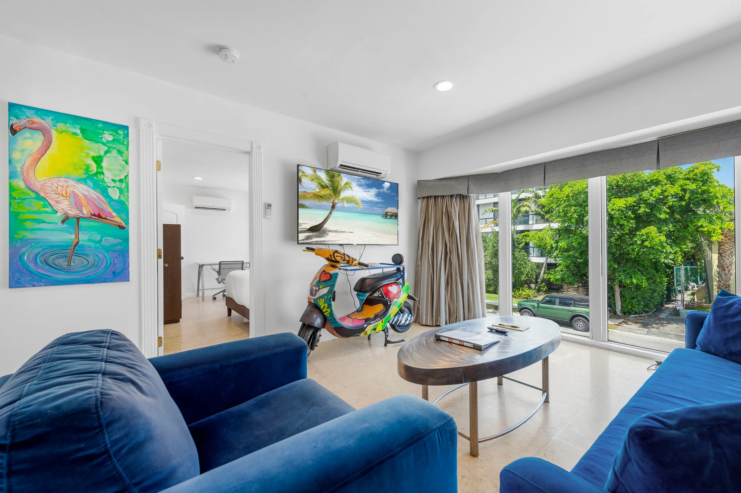 Bright living room with large windows, blue sofas, a colorful scooter, flamingo painting, and wall TV showing a beach scene.