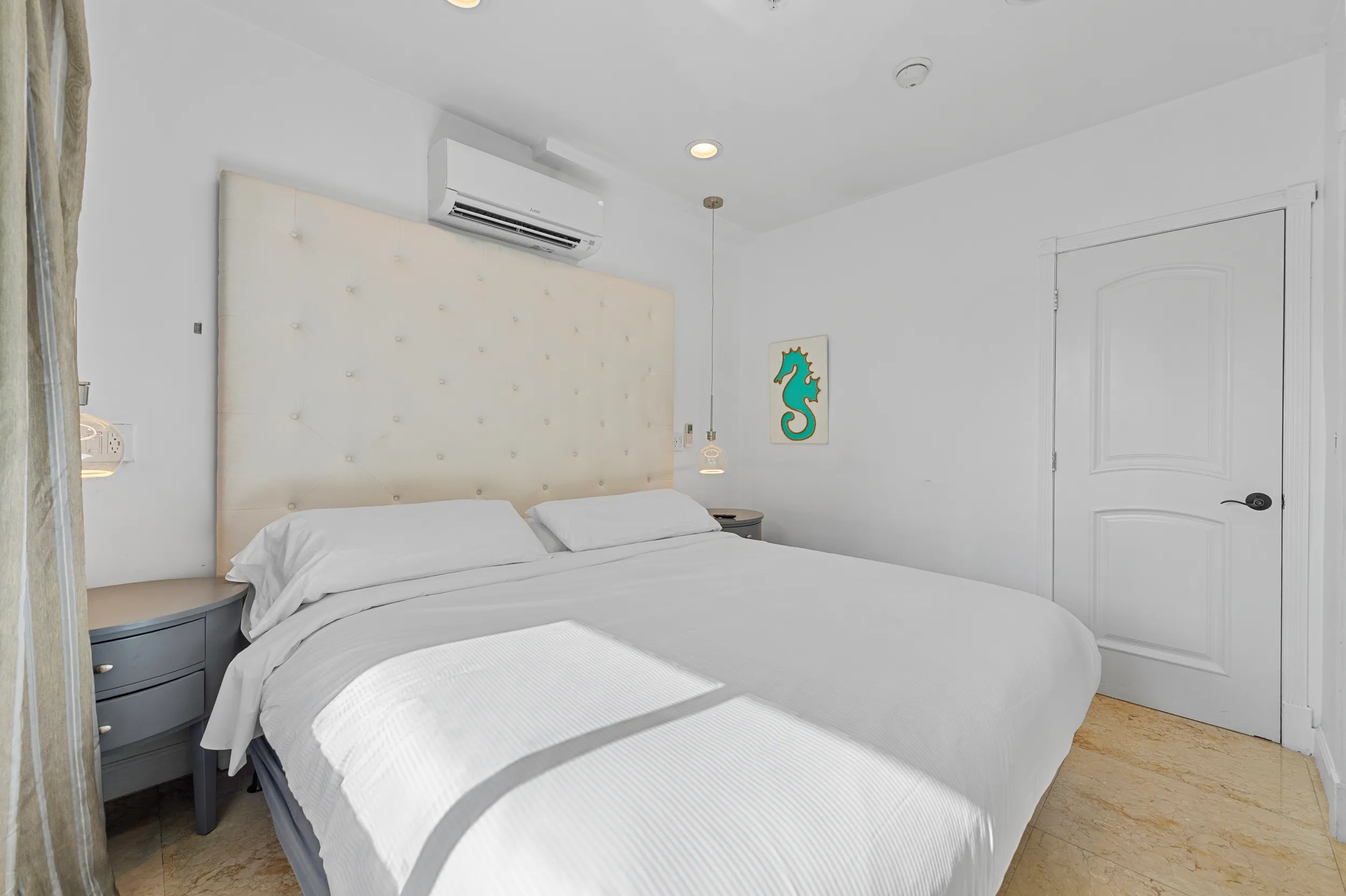 Bright bedroom with a large white bed, tufted headboard, bedside tables, and wall-mounted air conditioner.
