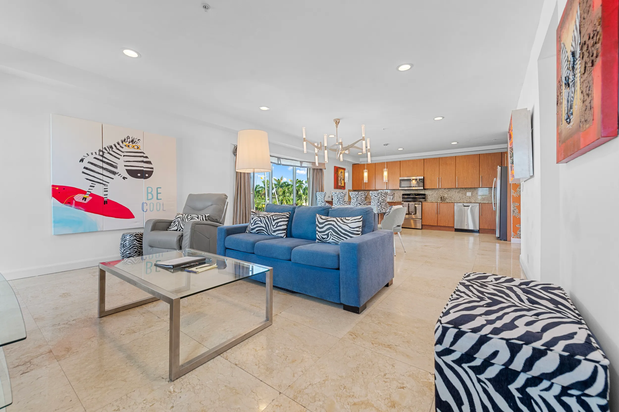 Modern living room with blue sofa, zebra-patterned cushions and ottoman, gray recliner, glass coffee table, and open kitchen with wooden cabinets.