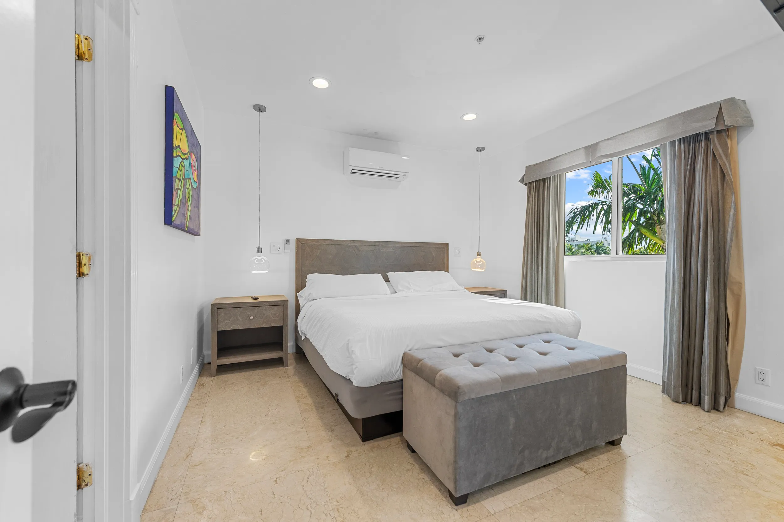 Minimalist bedroom with a king-size bed, white bedding, two wooden nightstands with hanging pendant lights, and a window showing palm trees outside.