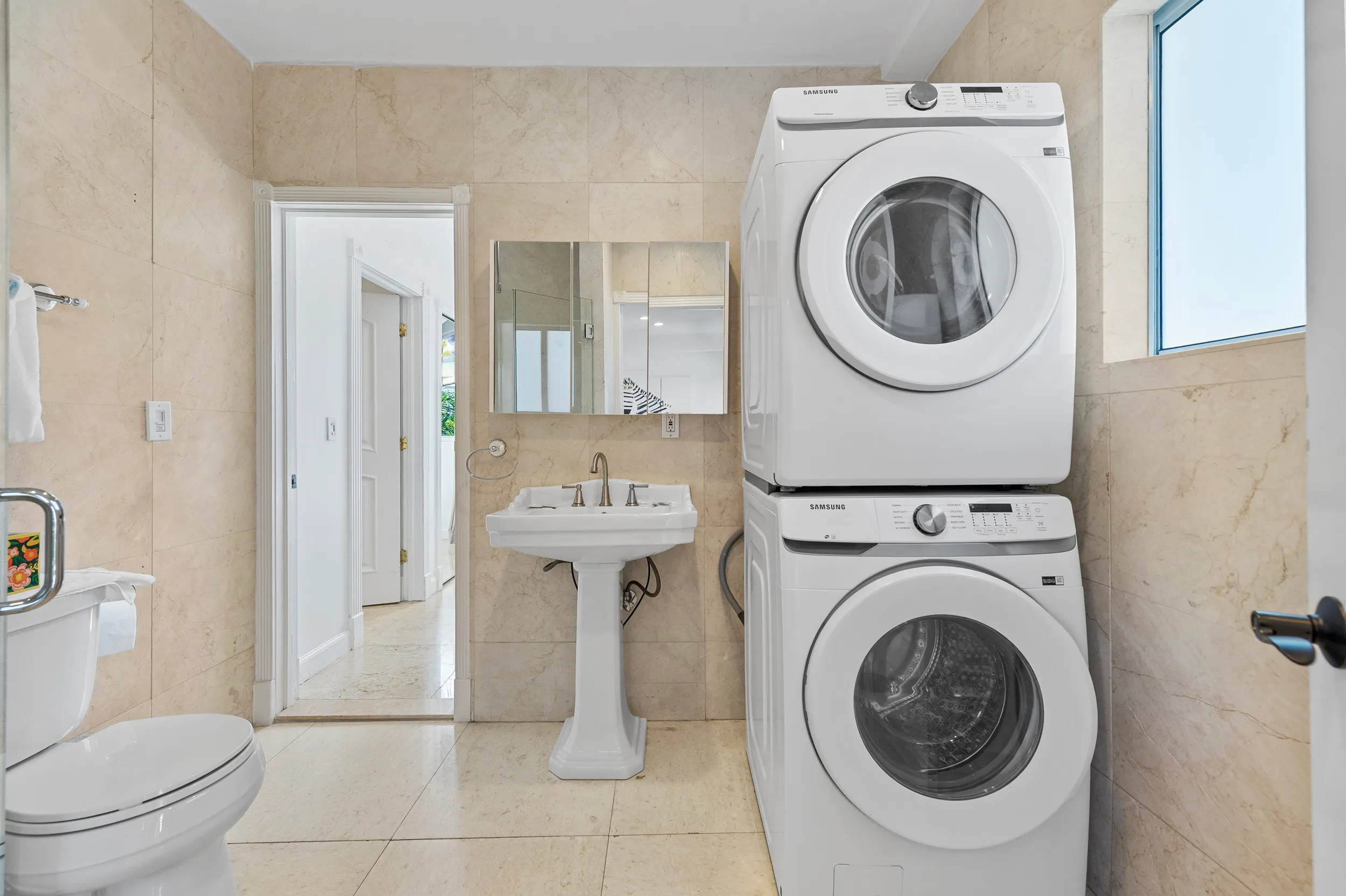 Bathroom with beige tiled walls and floor, a stacked Samsung washer and dryer unit, a white pedestal sink with a mirrored cabinet above, and a toilet on the left.