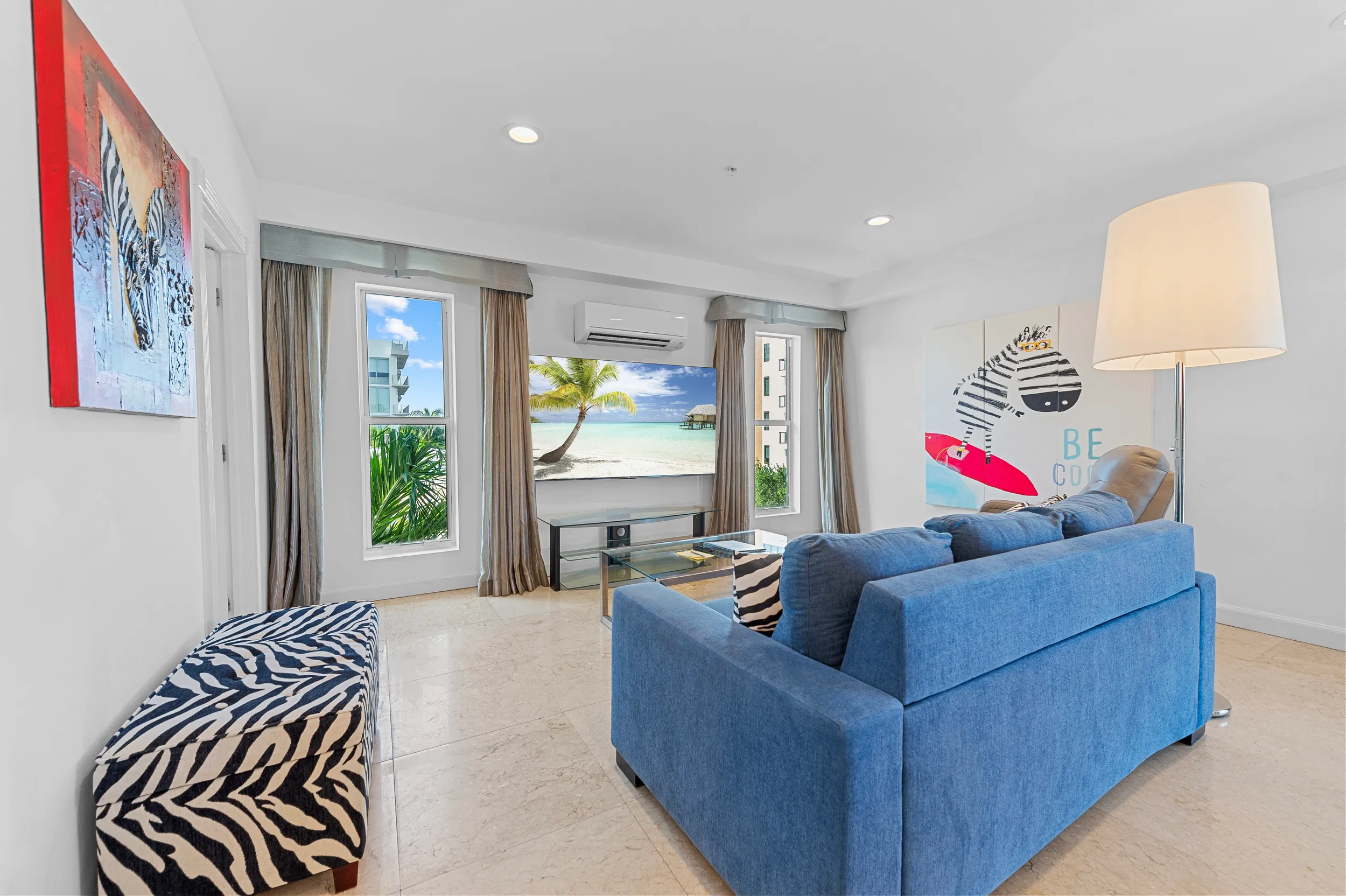 Modern living room with blue sofa, zebra-patterned bench and pillows, large floor lamp, tropical beach scene on TV, and zebra-themed wall art.