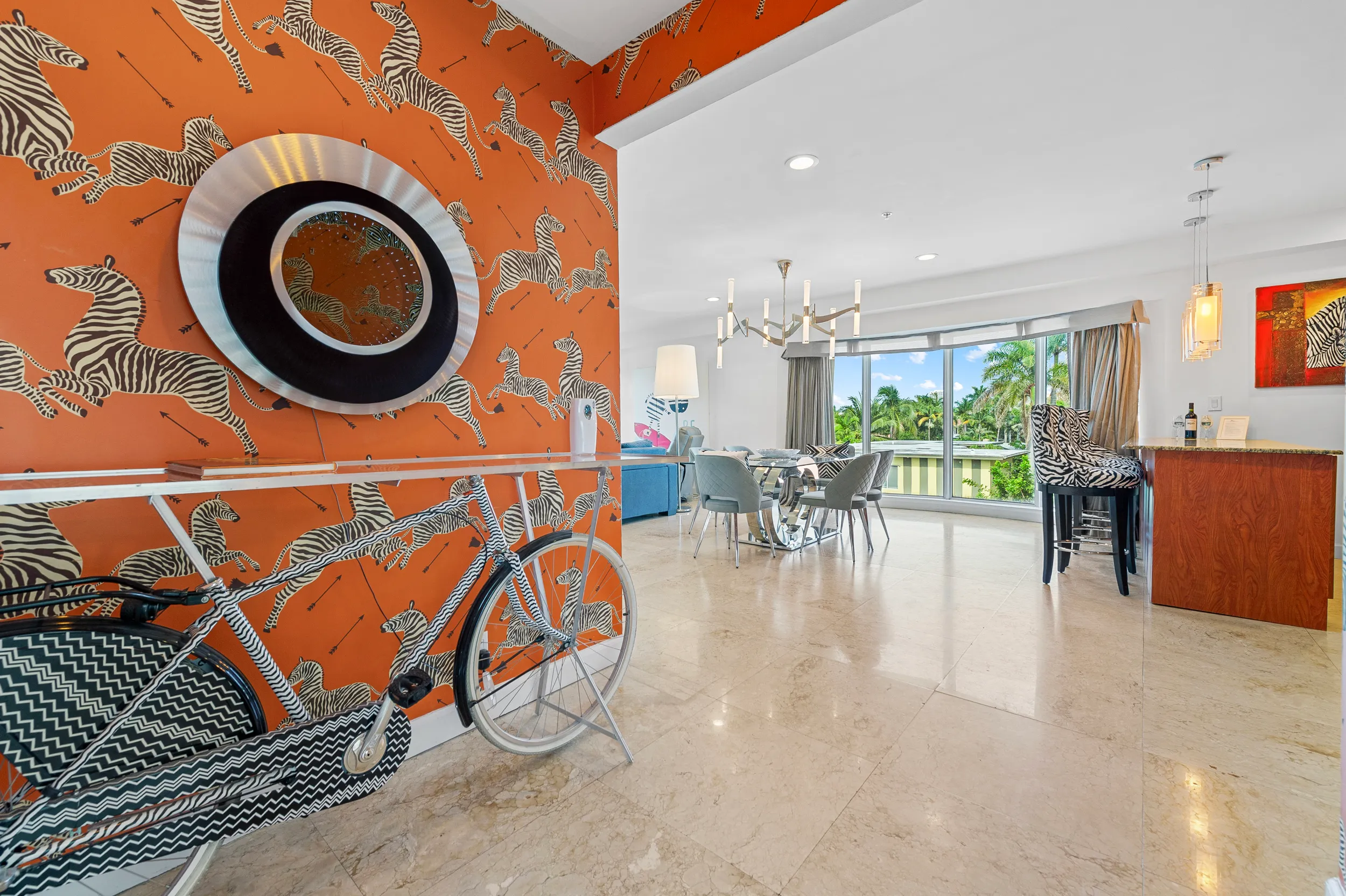 Modern living room with an orange zebra-patterned wall, a matching zebra-striped bicycle, glass dining table with chairs, bar area with zebra-bar stools, and large windows revealing palm trees outside.