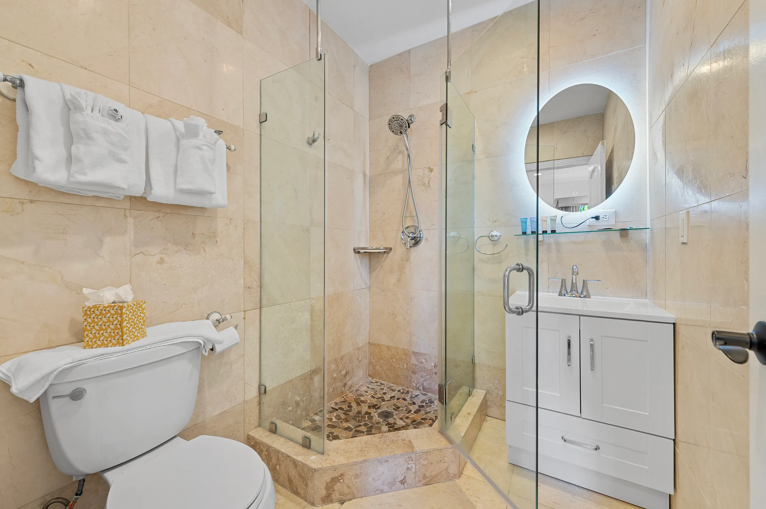 Modern bathroom with beige tiled walls, white toilet, glass-enclosed shower with pebble floor, and white vanity with illuminated round mirror.
