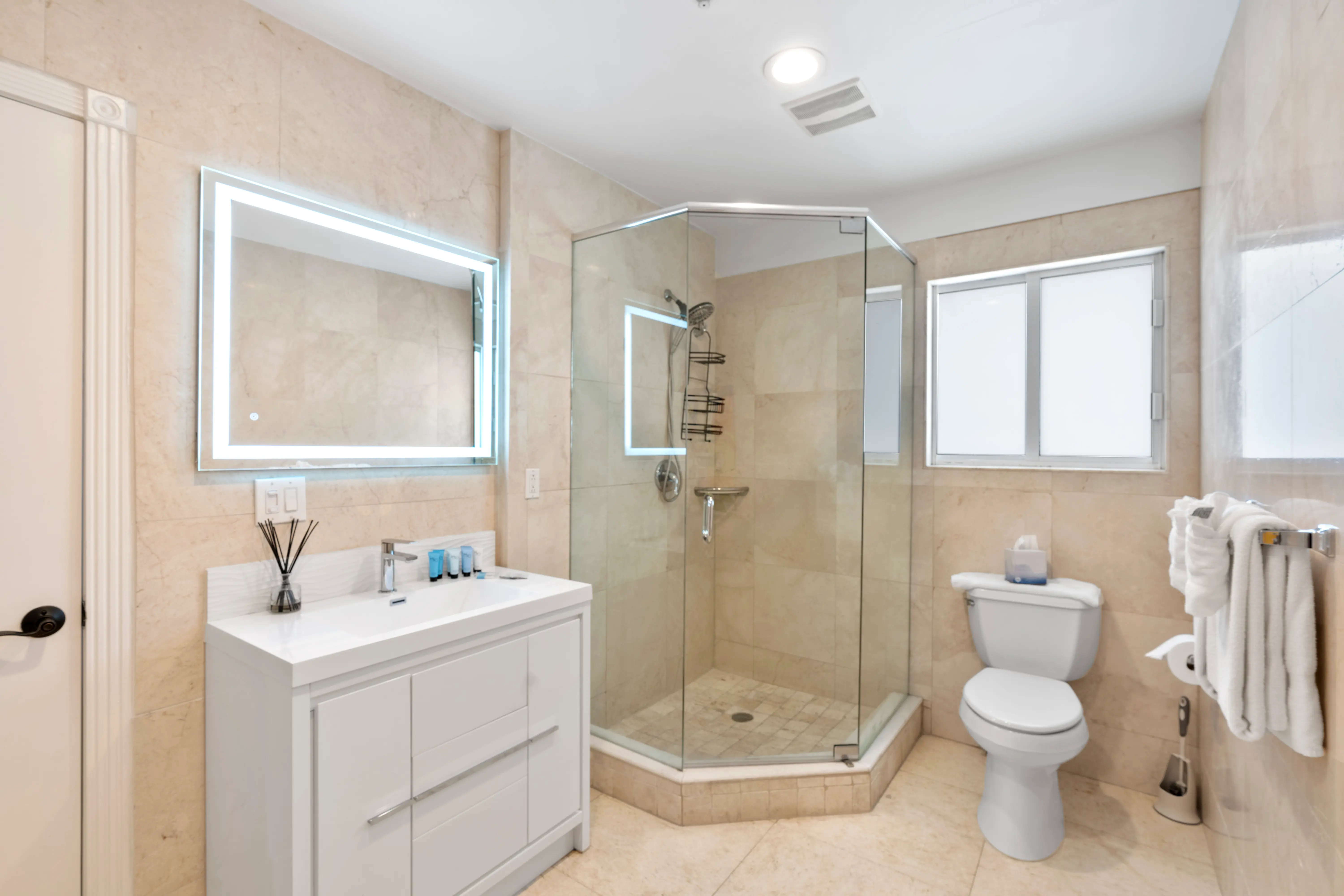 Modern bathroom with beige tiles, glass-enclosed corner shower, illuminated rectangular mirror, white vanity, and toilet beside a frosted window.