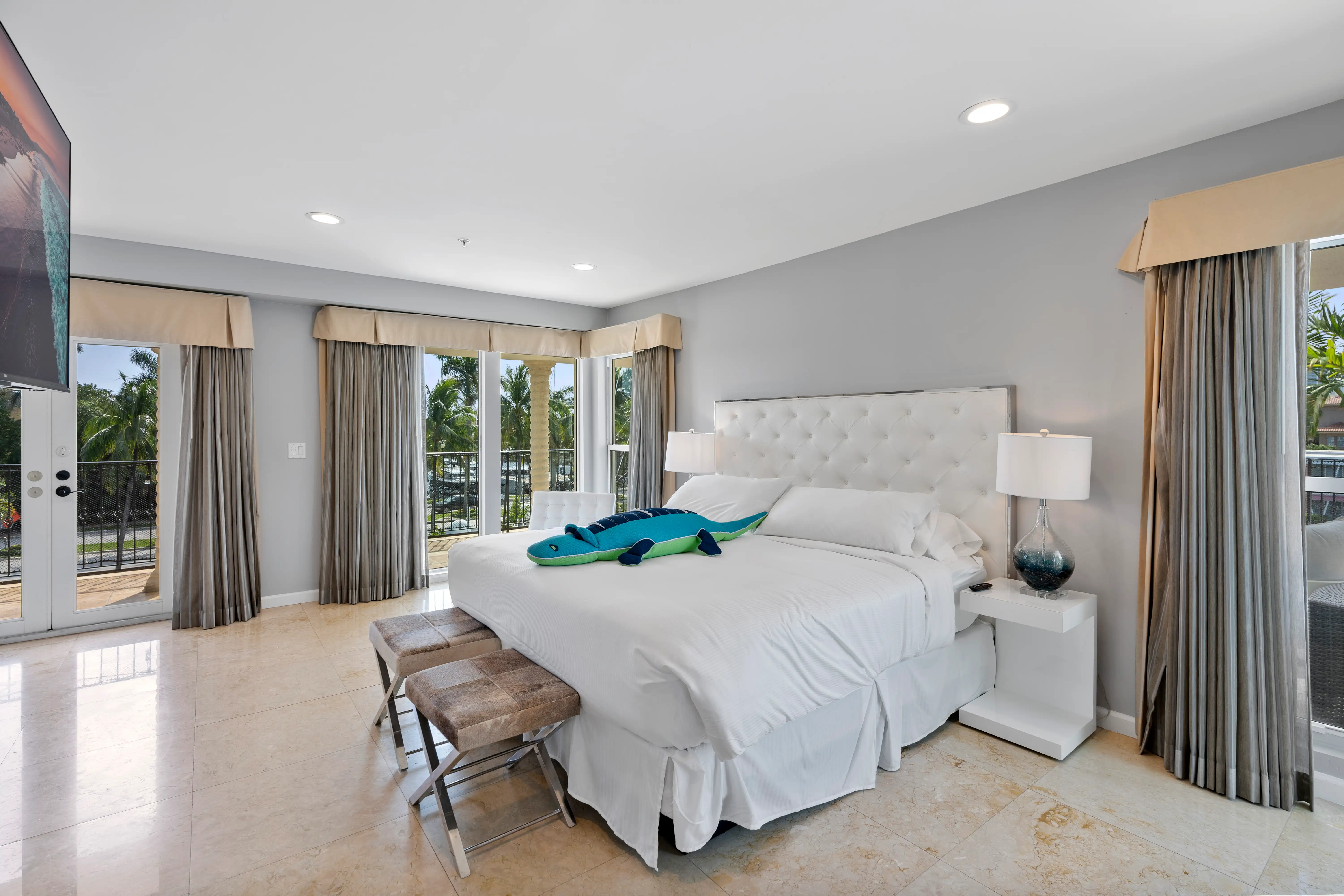 Modern bedroom with large white bed, two stools at foot, glass doors opening to balcony with palm trees outside.