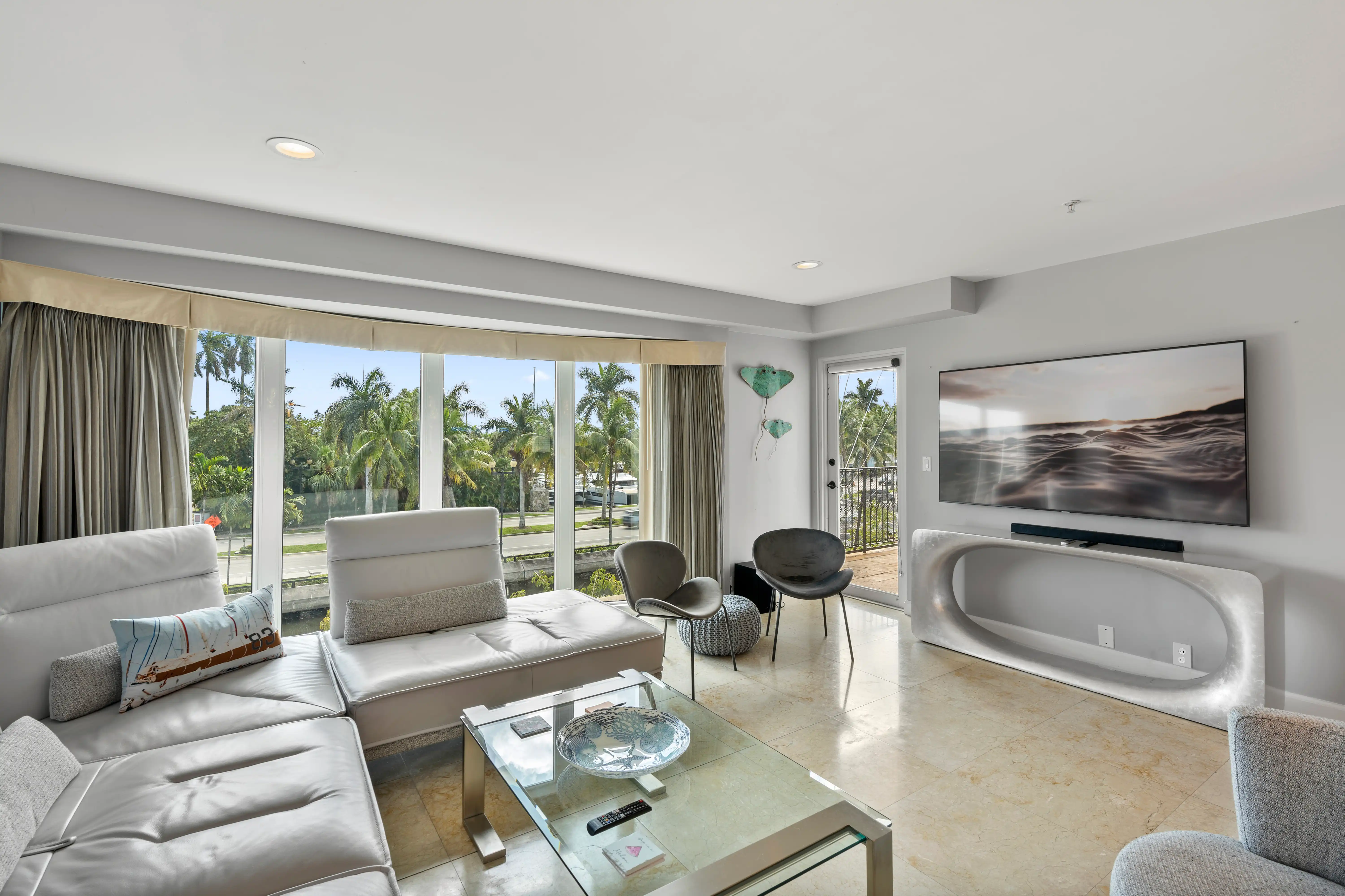 Modern living room with white sectional sofa, glass coffee table, two gray chairs, mounted flat-screen TV, and large windows showing palm trees outside.