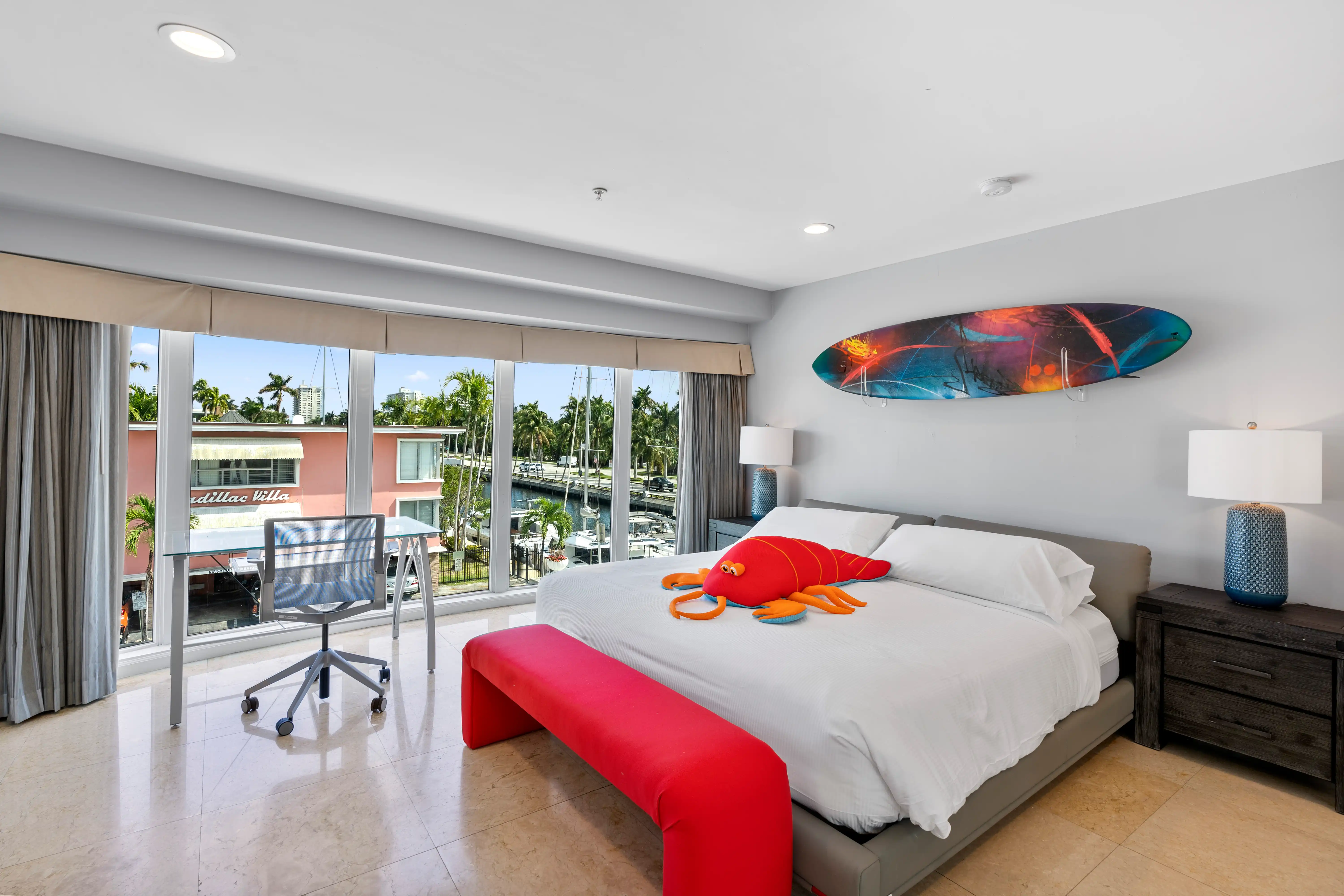 Modern bedroom with large windows overlooking marina, a king bed with white bedding and red lobster plush toy, red bench at foot of bed, desk with chair by the window, and surfboard wall art.