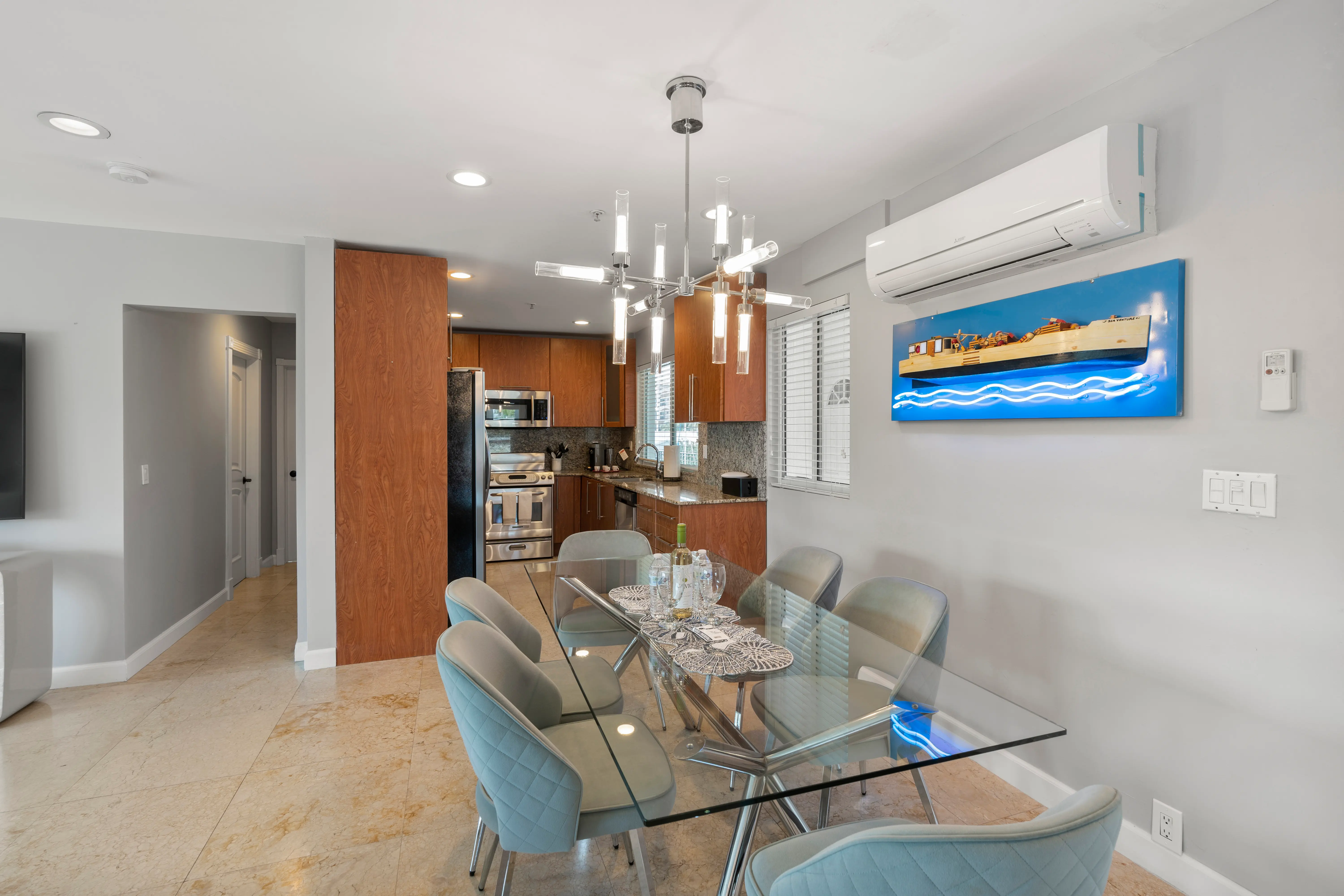 Modern dining area with glass table, six upholstered chairs, chandelier, and kitchen with wooden cabinets in the background.