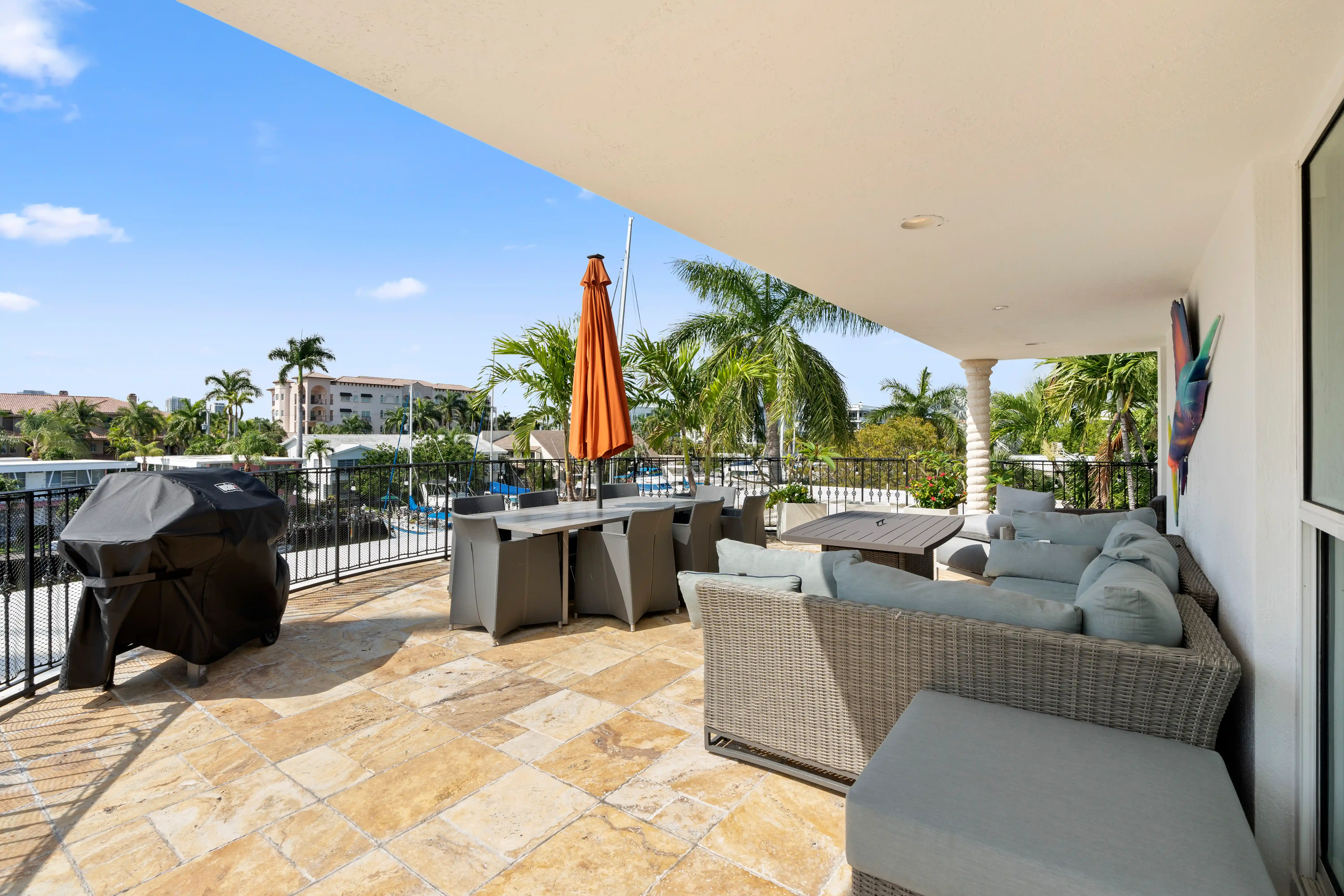 Spacious outdoor patio with beige tiled floor, wicker sectional sofa with light cushions, dining table with chairs and orange umbrella, and a covered grill, overlooking palm trees and nearby buildings.