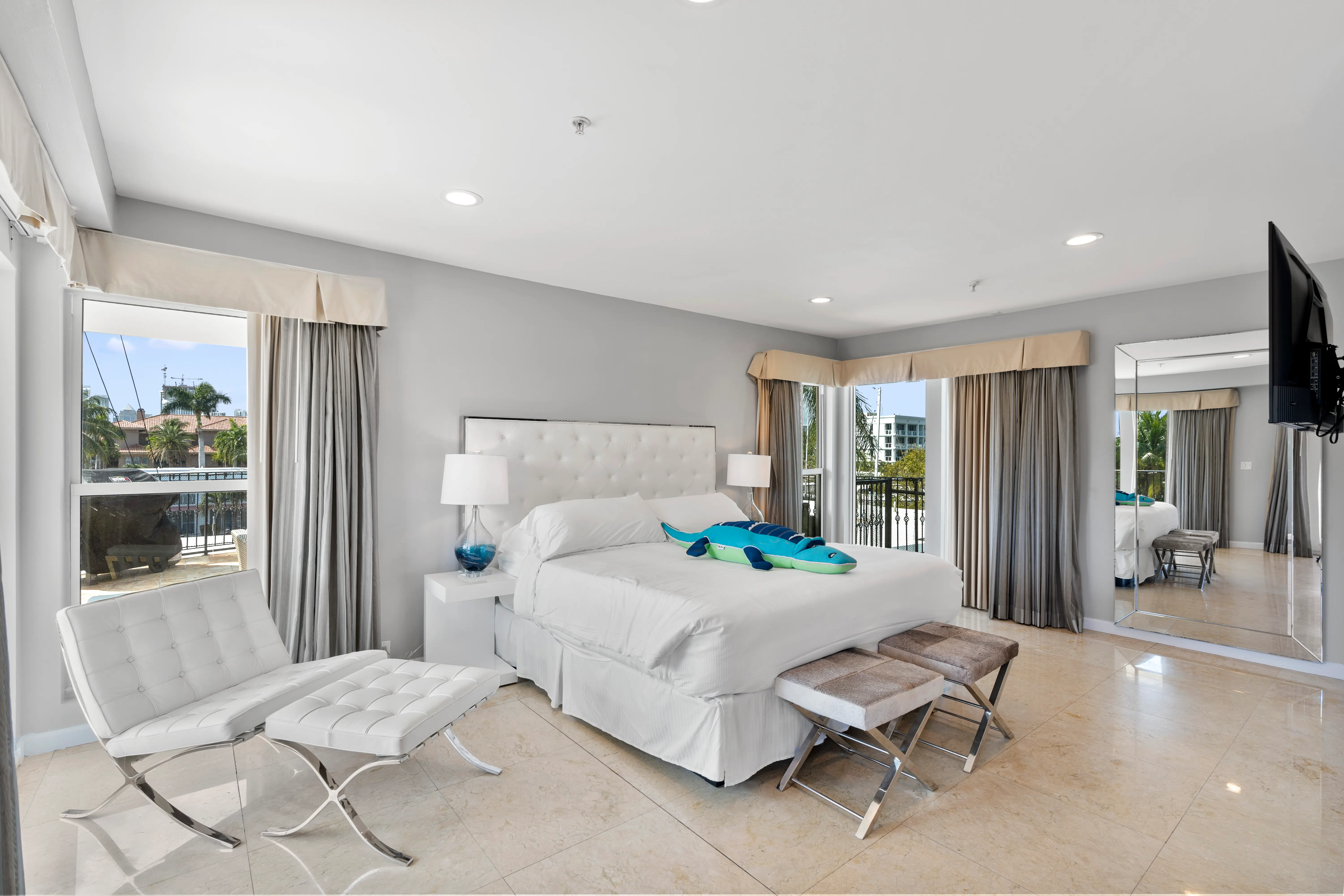 Bright hotel room with a large white bed, two stools, a white chair with footrest, nightstands with lamps, a wall-mounted TV, and two windows with curtains showing outdoor greenery.