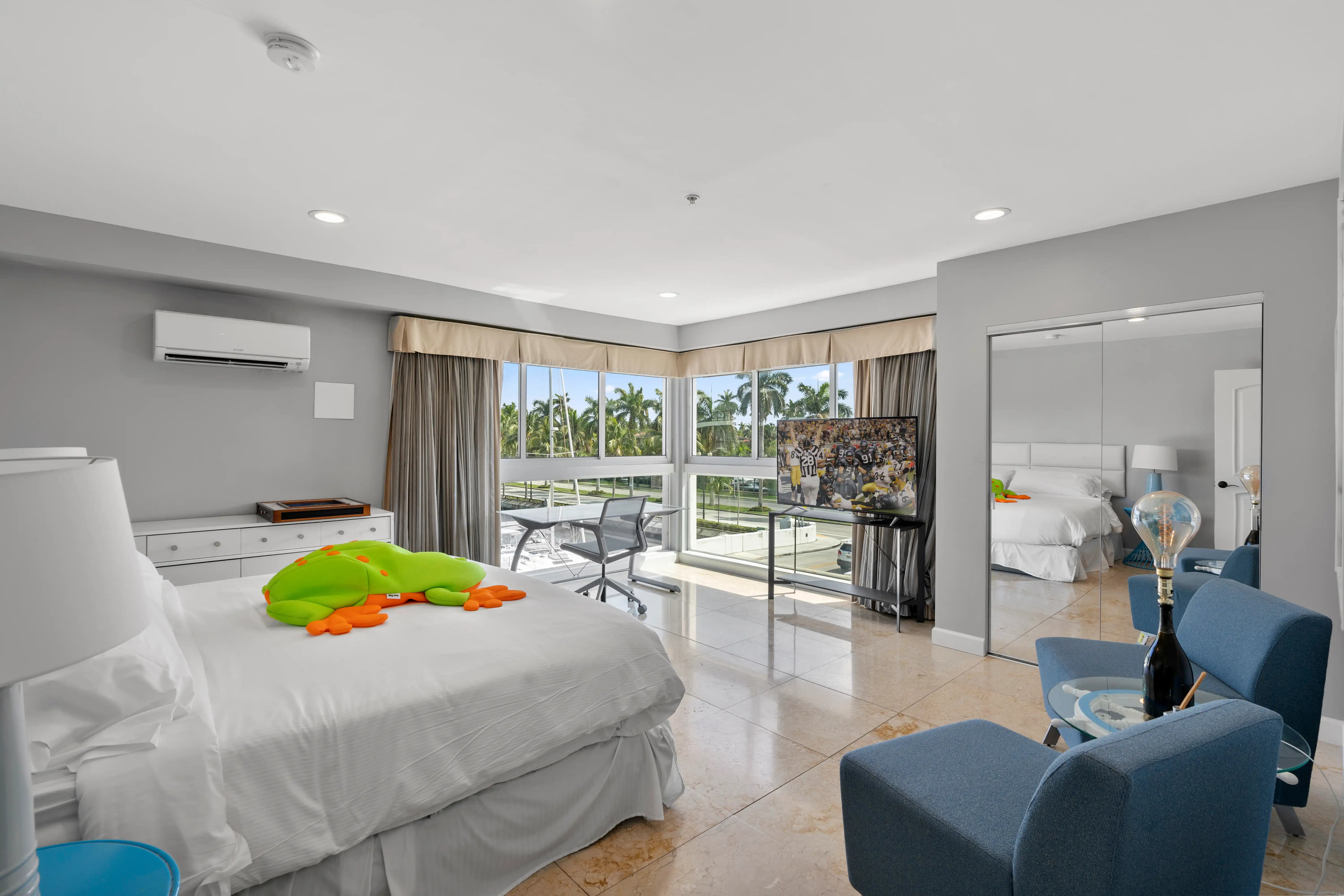 Bright modern bedroom with a large bed topped with a green and orange stuffed frog, corner windows with palm tree view, flat-screen TV, blue armchairs, and mirrored closet doors.