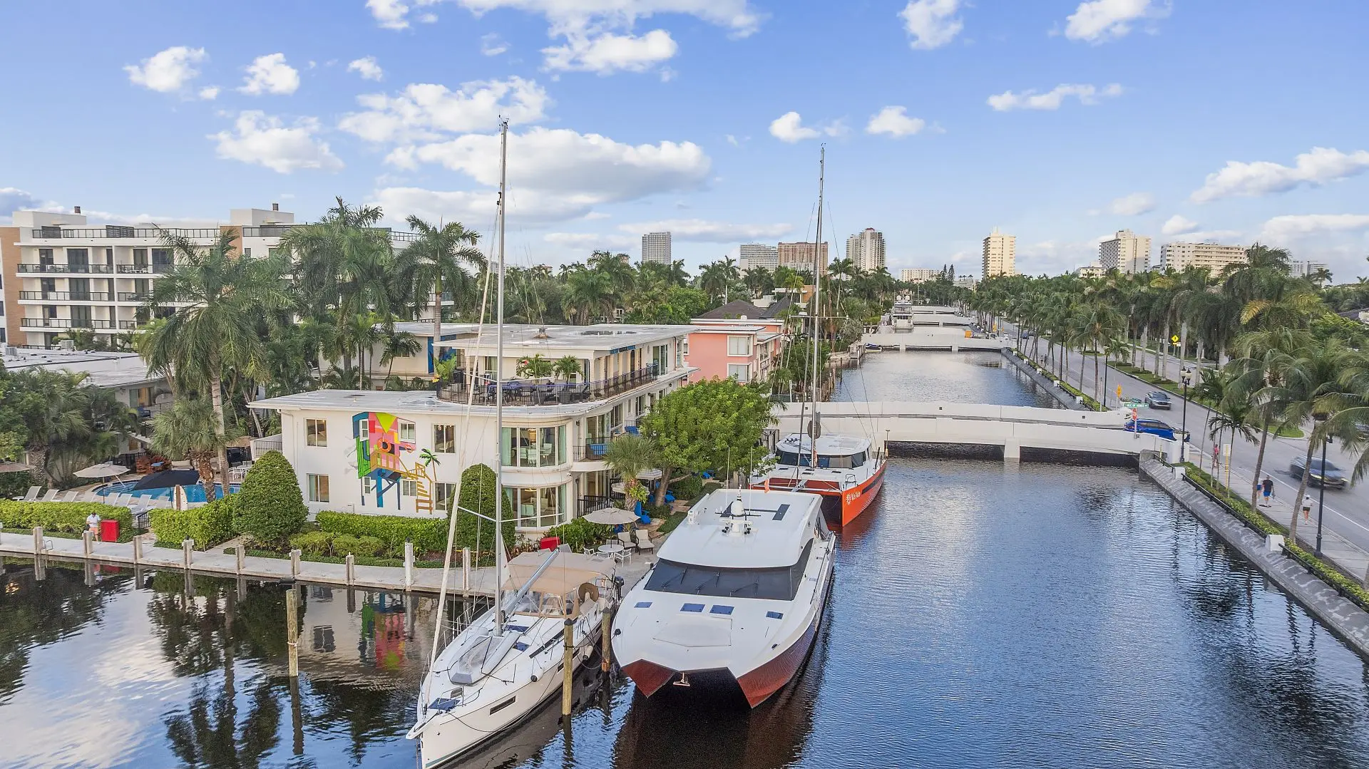 Boats docked along a canal lined with palm trees and buildings under a partly cloudy blue sky.