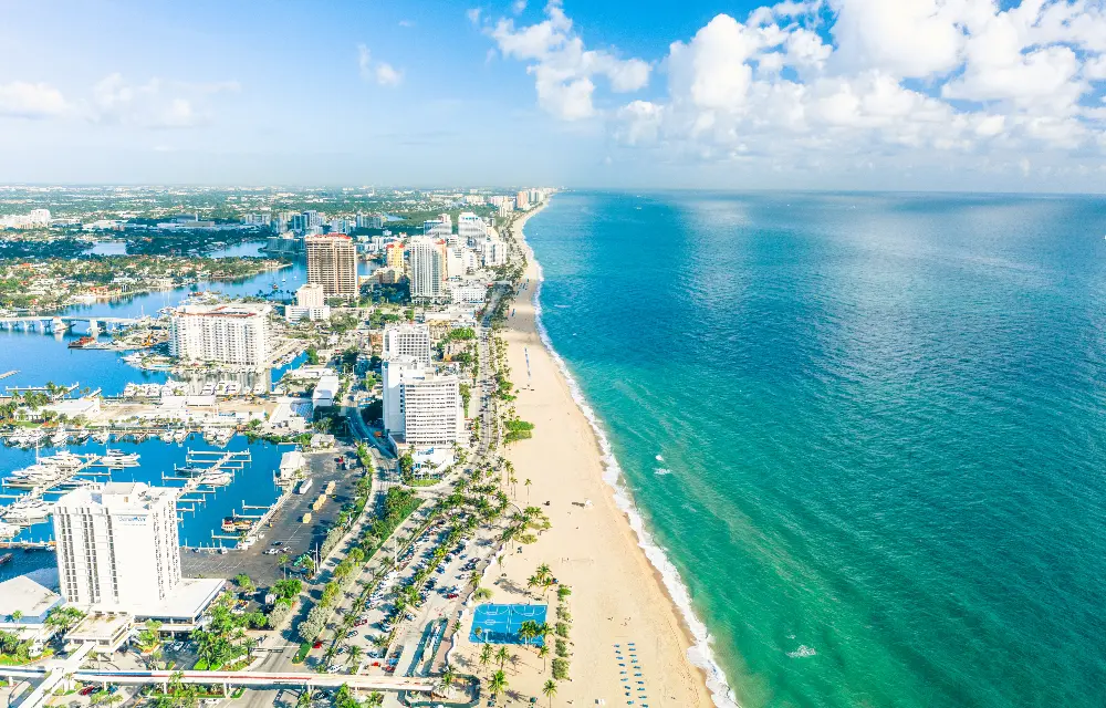 Aerial view of a coastal city with a long sandy beach, turquoise ocean, palm trees, and high-rise buildings along the shoreline under a partly cloudy sky.