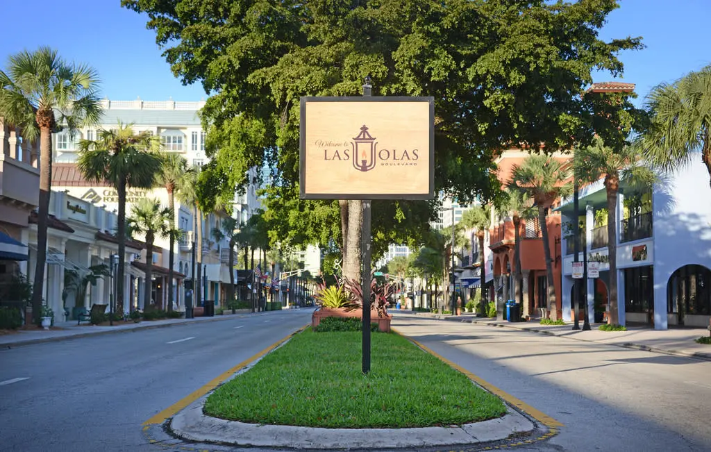 Welcome sign for Las Olas Boulevard centered on a grassy median with palm trees and buildings lining both sides of a sunny street.