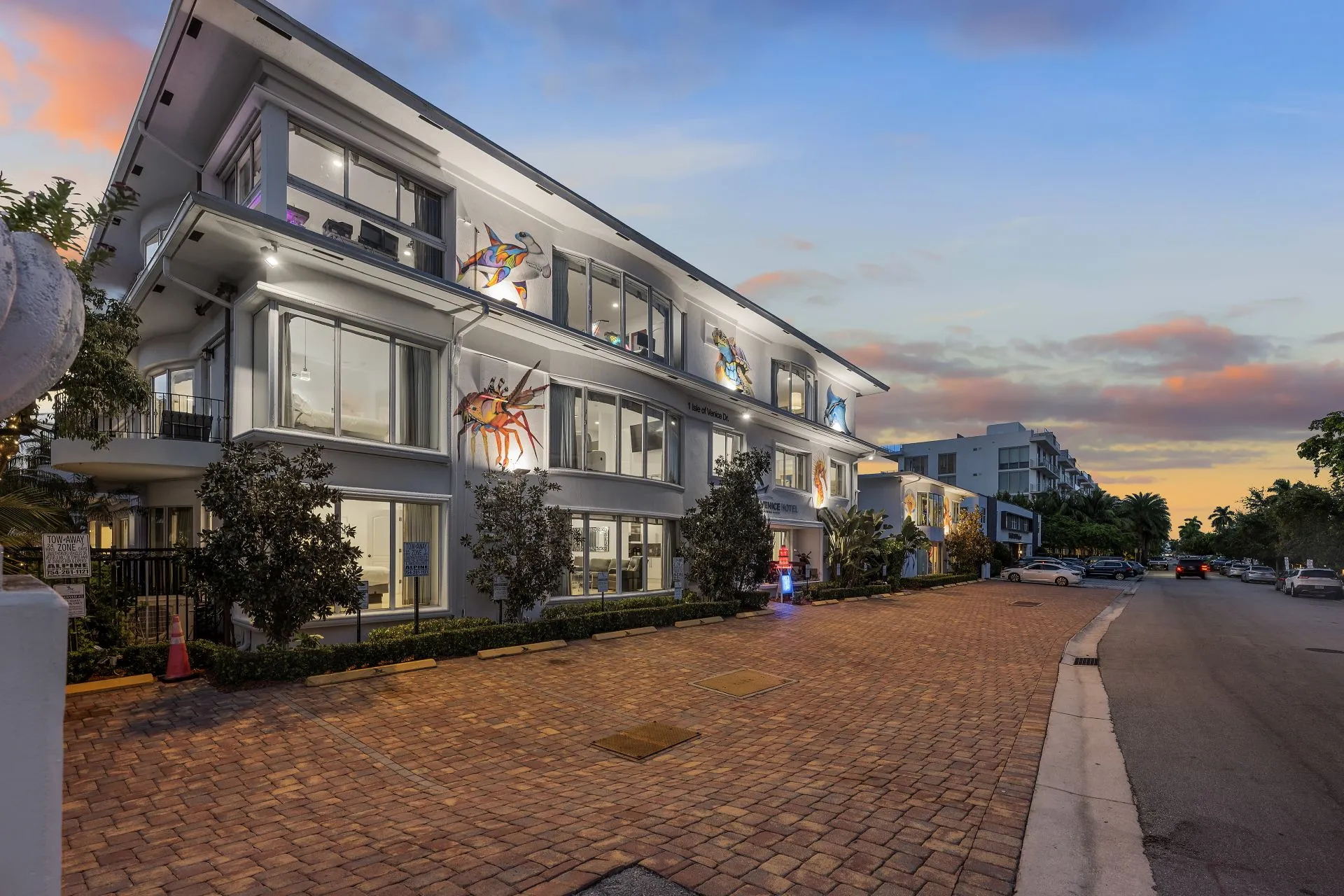 White multi-story building with large windows and colorful marine animal murals on the walls by a brick-paved parking area at sunset.